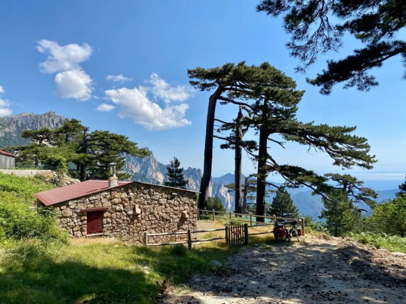 Stone refuge with red roof, pine trees, and rugged mountains near the sea, Col de Bavella.