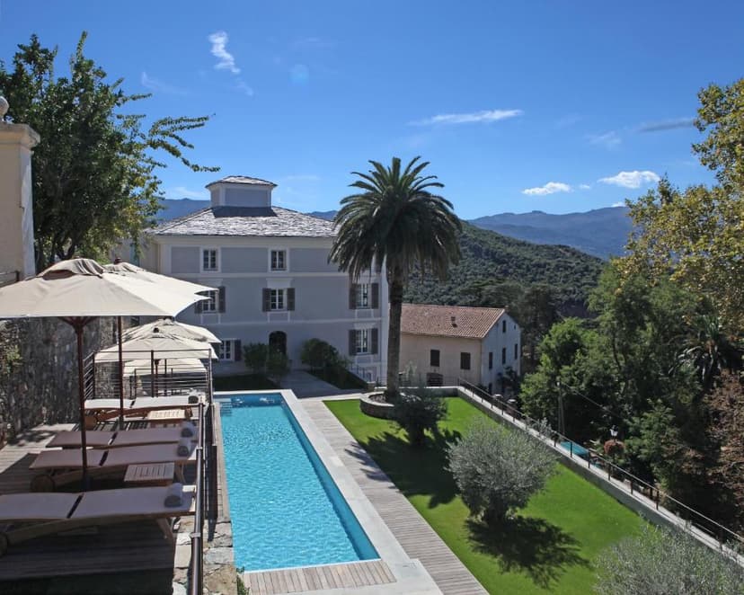 Swimming pool and sun loungers by a historic building with mountains in Corsica.