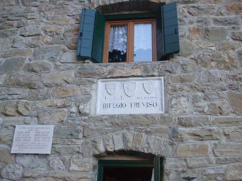 Stone wall entrance with "Rifugio Treviso" plaque, window with green shutters, and memorial plaque.