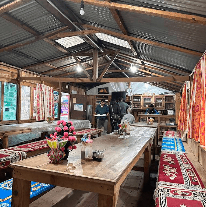 Rustic wooden dining hall interior with long tables, colorful rugs on benches, and staff serving food.