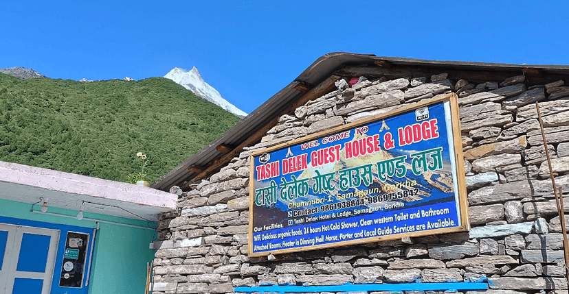 Tashi Delek Guest House & Lodge sign on stone wall with green mountain and snowy peak background.