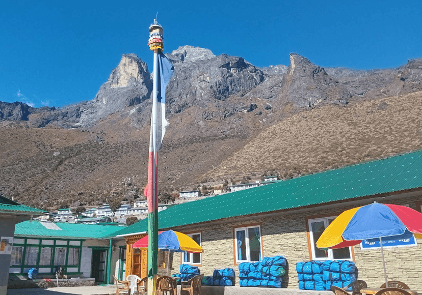 Lodge exterior with prayer flag pole, colorful umbrellas, and steep brown mountains under blue sky.