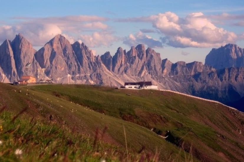 Alpine meadow with mountain huts below jagged peaks under a partly cloudy sky