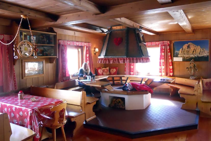 Interior of Rifugio Giussani mountain hut with wooden paneling, central stove, and woman seated by a window.