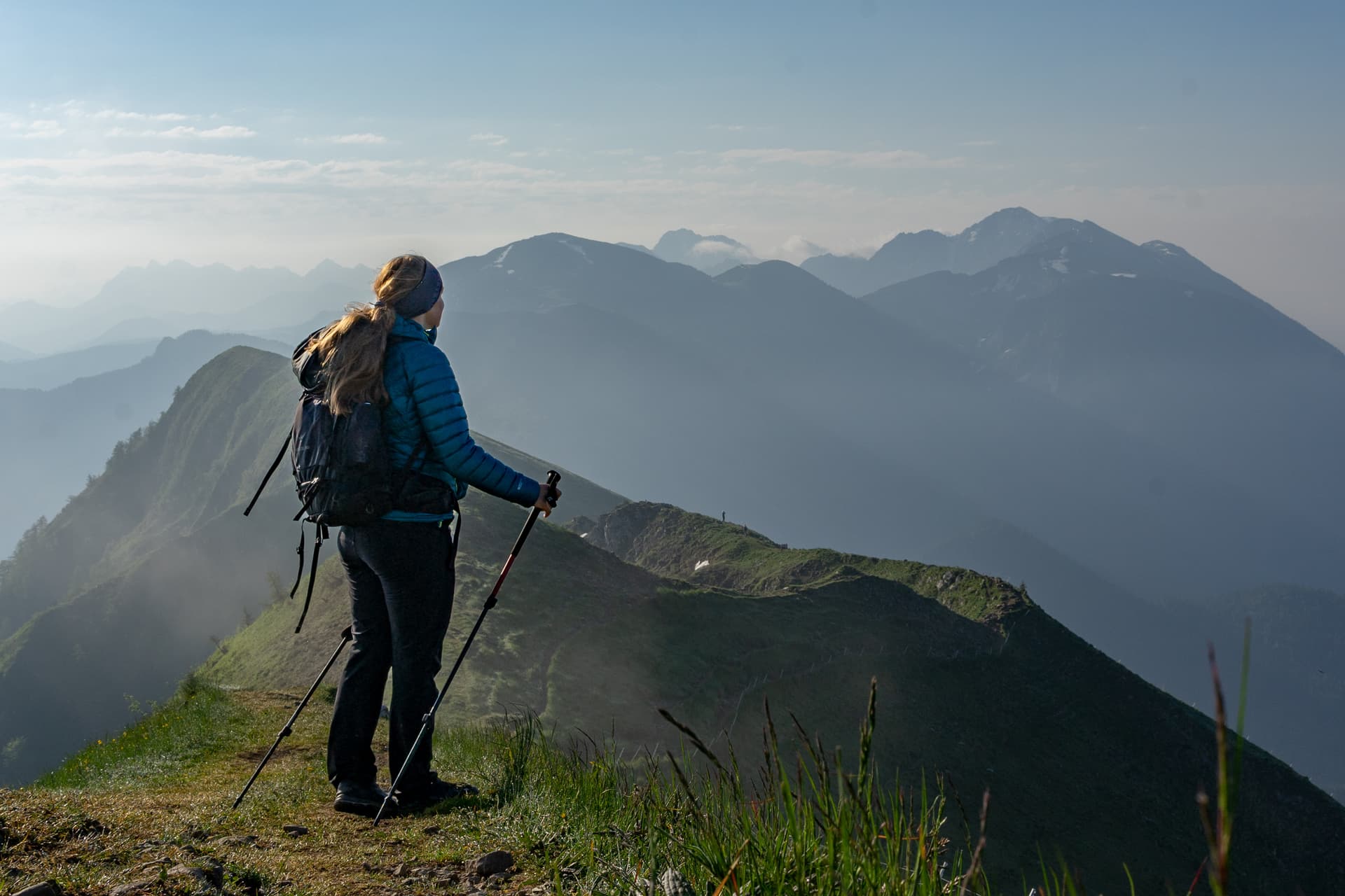 Hiker with backpack and poles standing on grassy ridge overlooking misty mountain range.