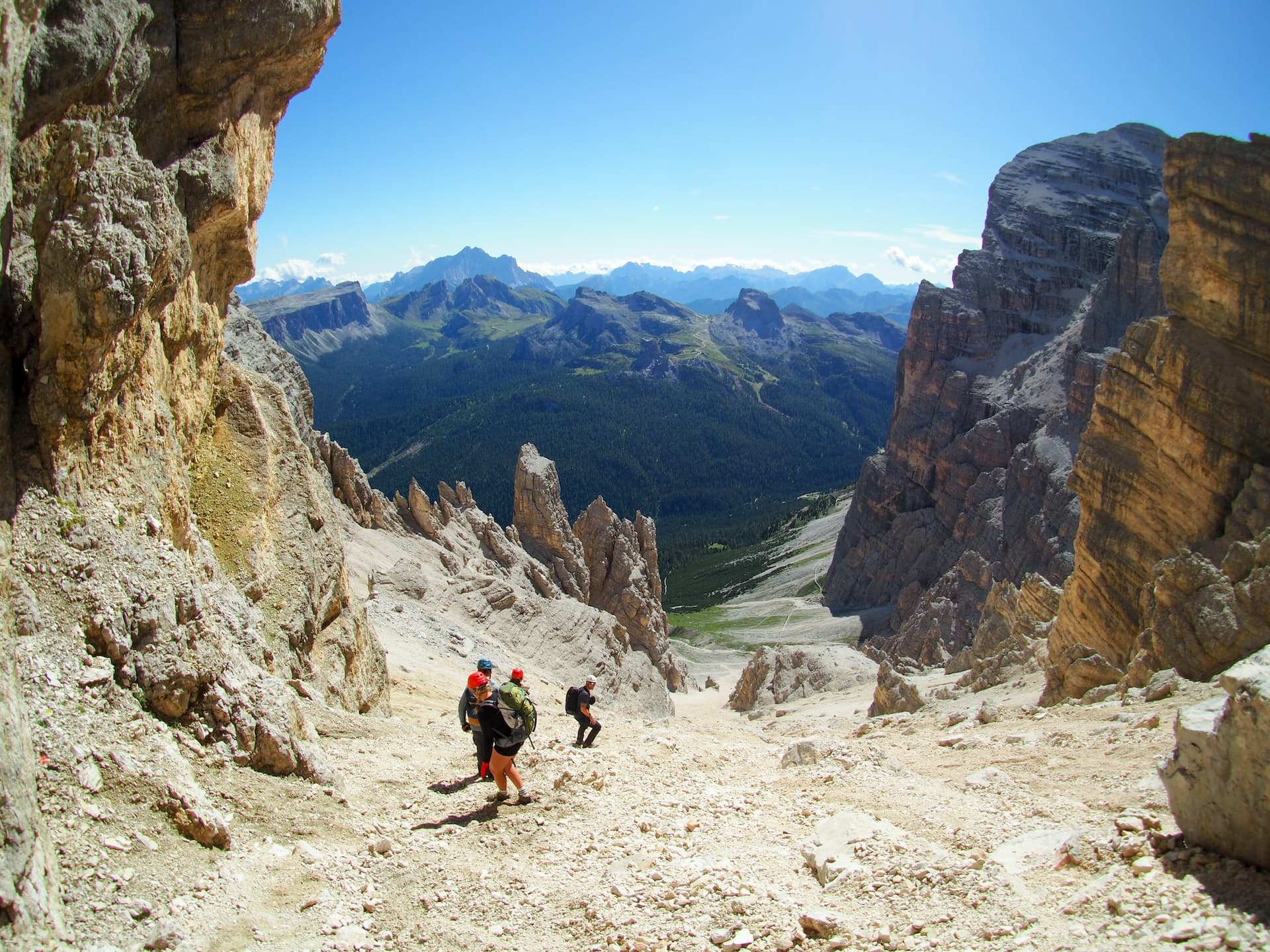 Hikers descending steep, rocky terrain between towering cliffs with a view of forested mountains.