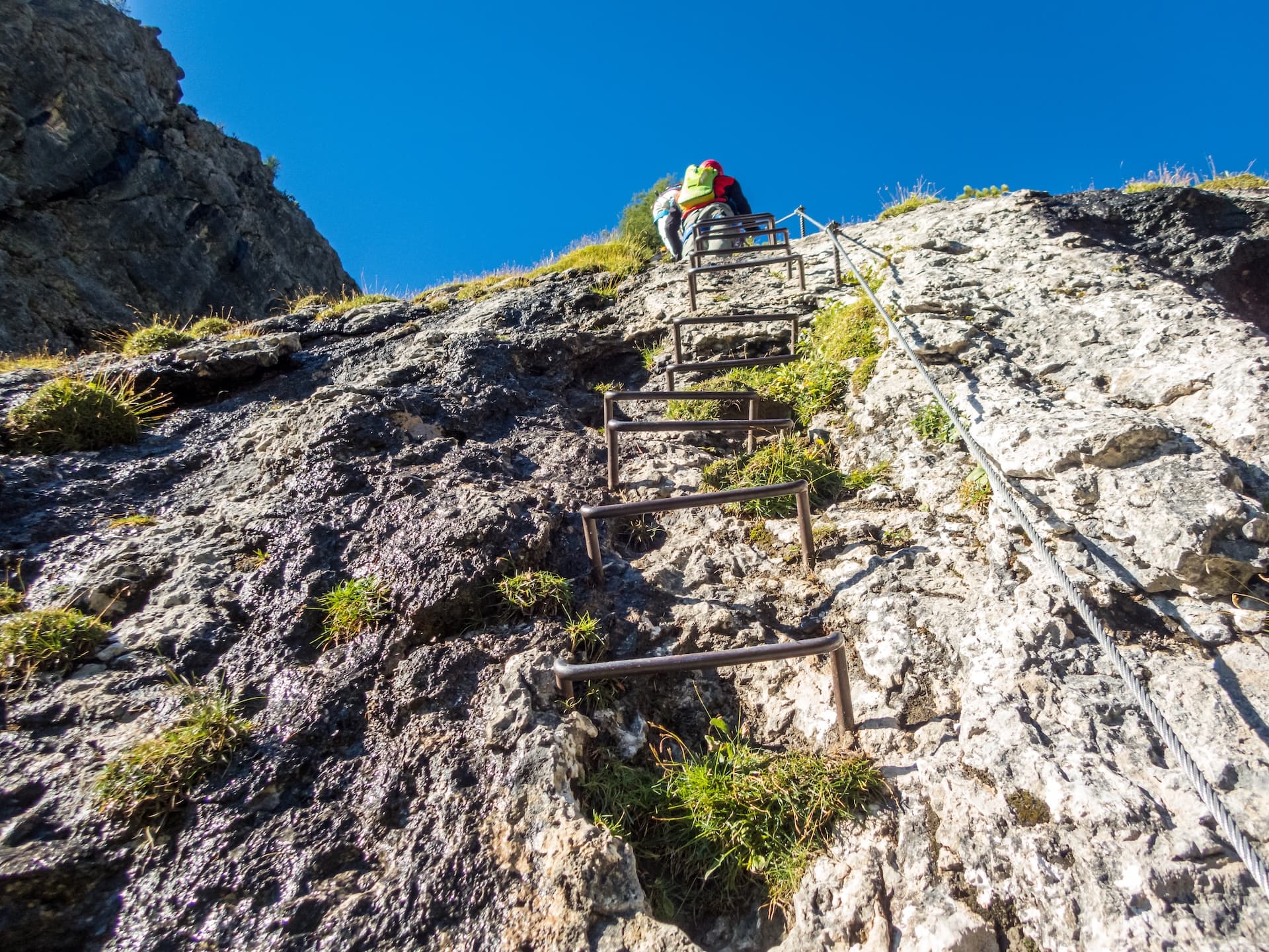 Hikers ascending steep rock face using metal rungs and cable on a via ferrata under blue sky.