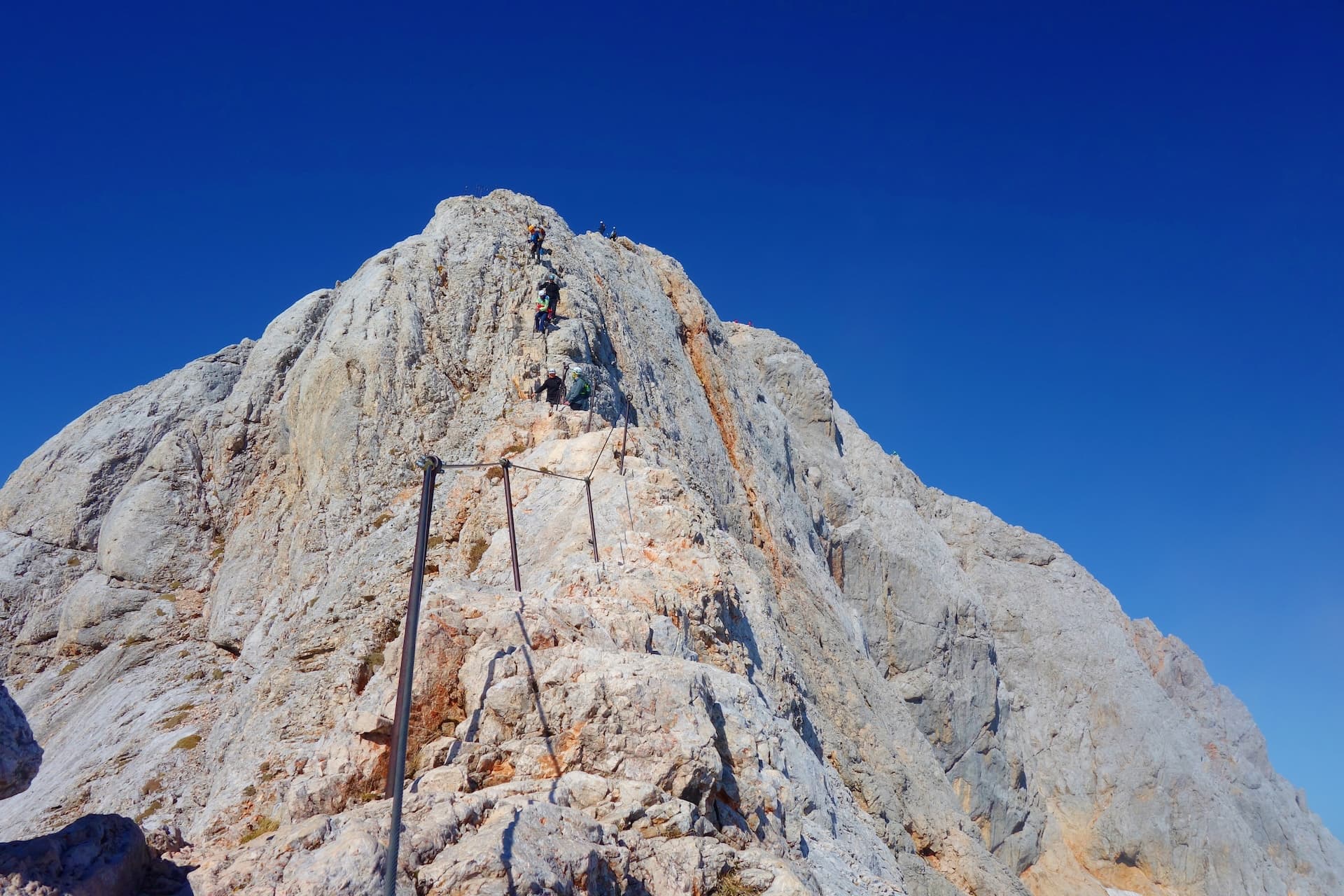 Hikers ascending steep, rocky mountain face with via ferrata cables under clear blue sky.