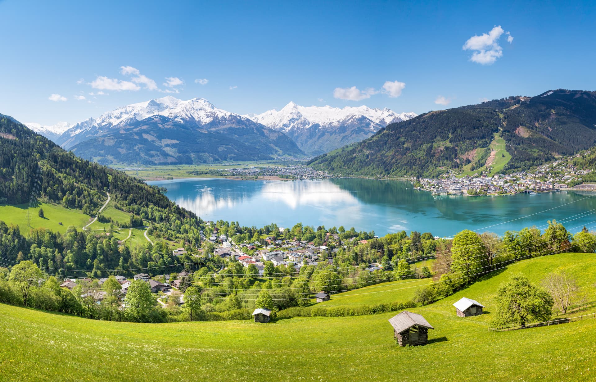 Alpine lake town nestled between green hills and snow-capped mountains under blue sky