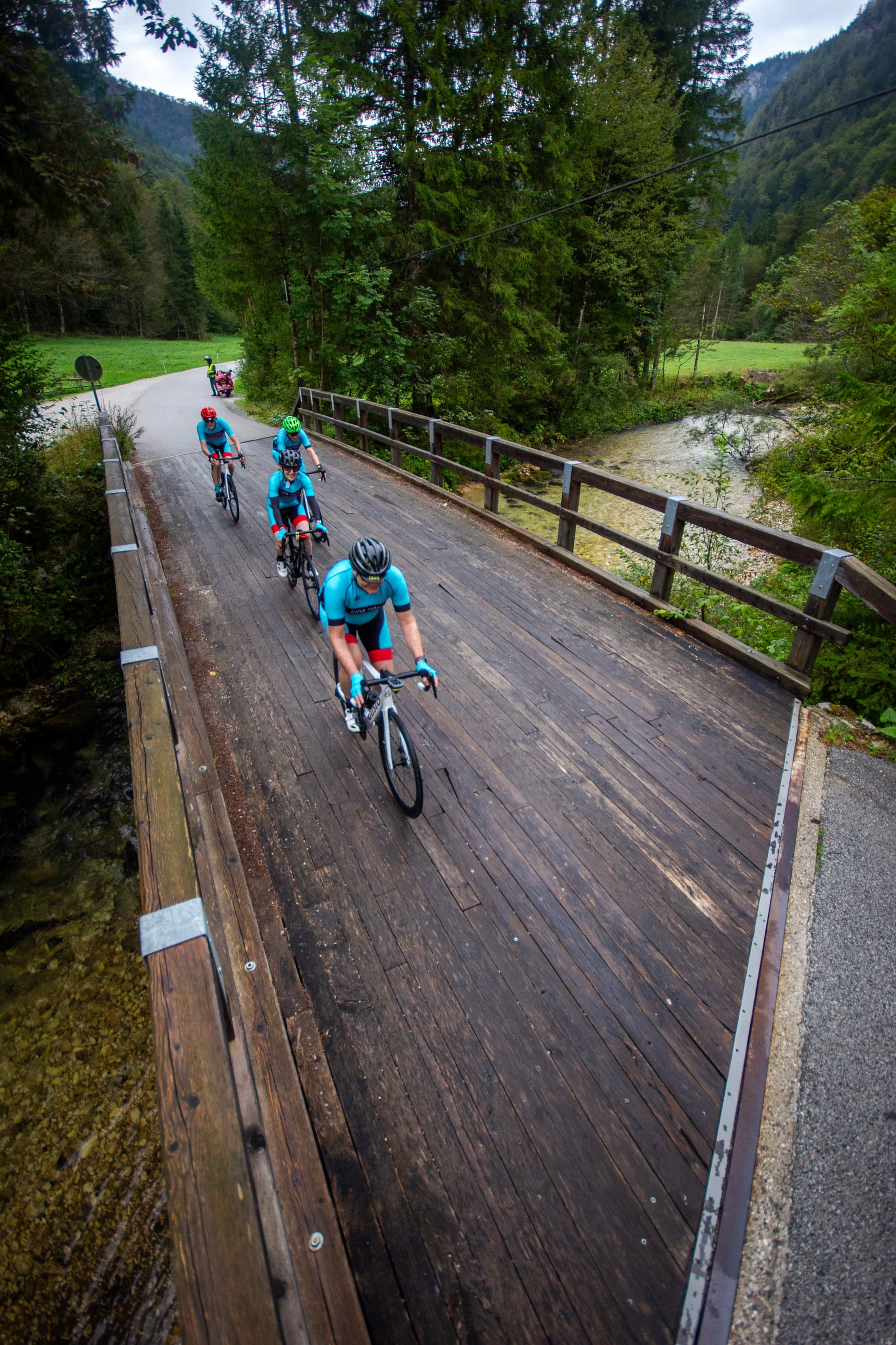 Cyclists in blue jerseys ride across a dark wooden bridge over a shallow stream in a forested mountain valley.