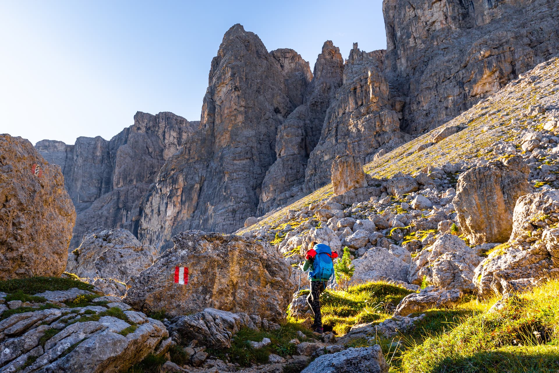 Hiker with blue backpack ascending rocky trail beneath towering gray mountain cliffs