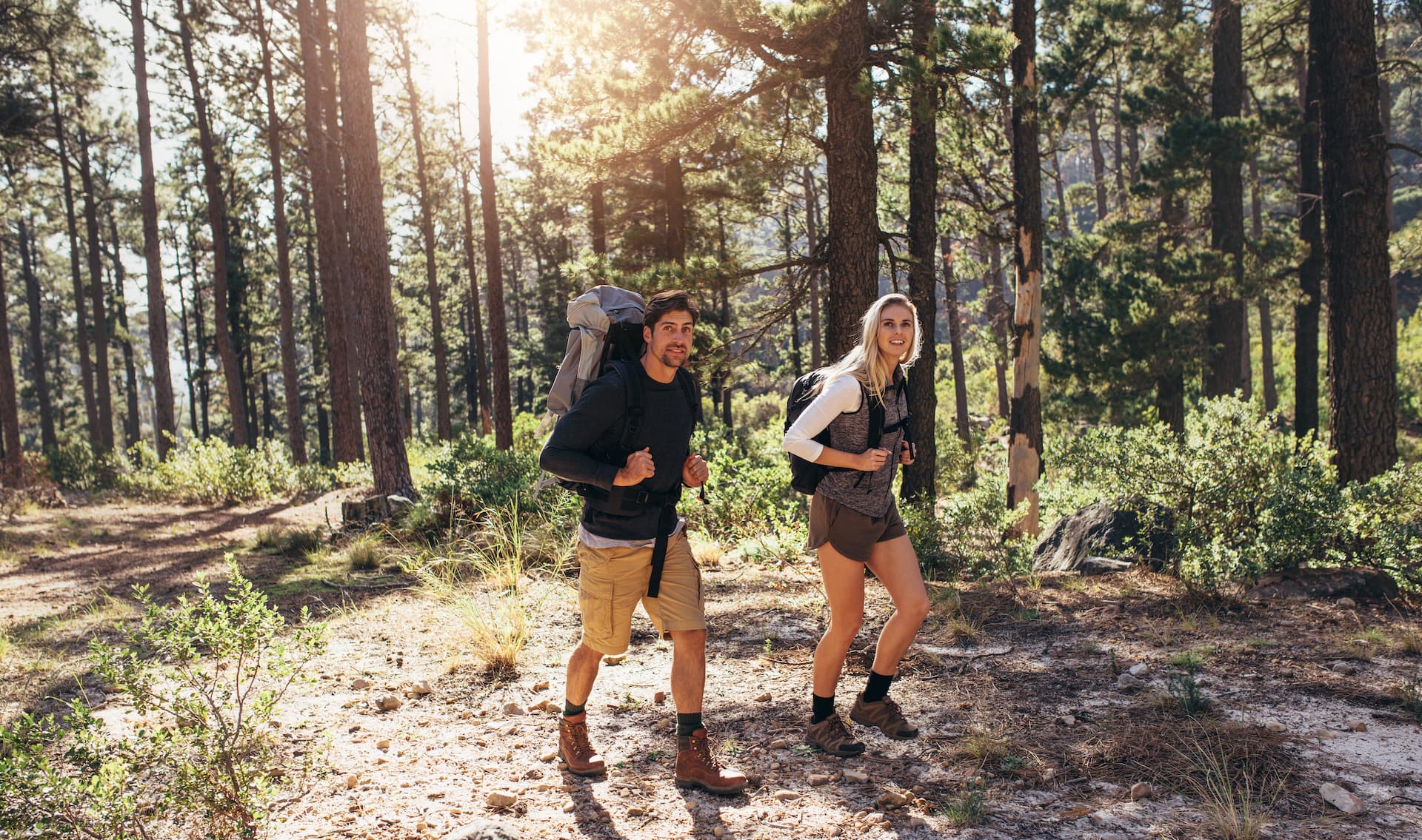 Couple hiking with backpacks on a sunny, rocky trail through a dense pine forest.