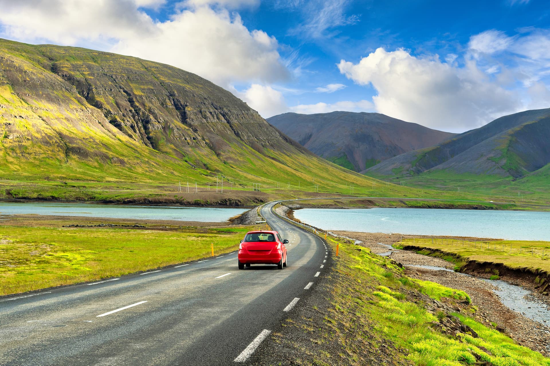 Red car driving on road between green mountains and blue water in Iceland