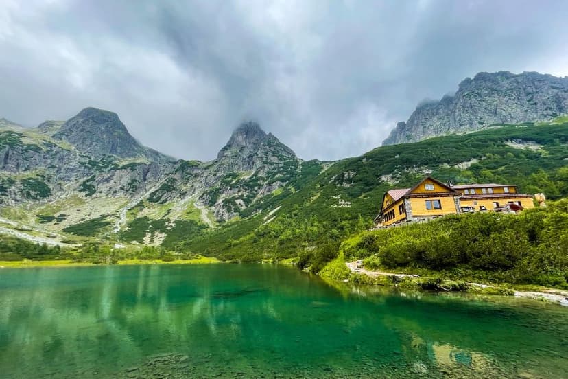Zelene Pleso Hut by emerald alpine lake with rugged mountains under cloudy sky.