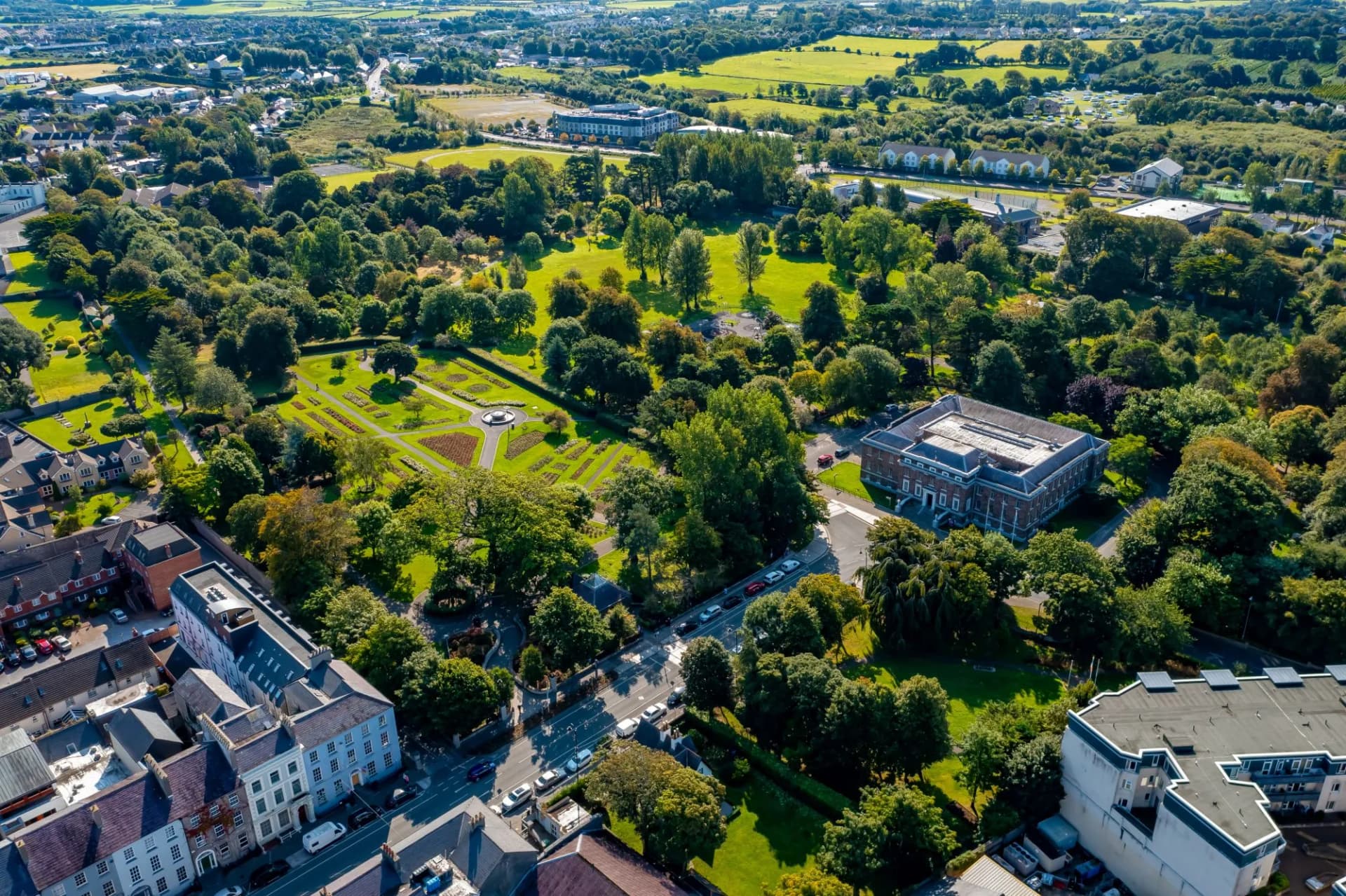 Aerial view of Tralee town park with formal gardens, dense trees, and surrounding buildings on a sunny day.