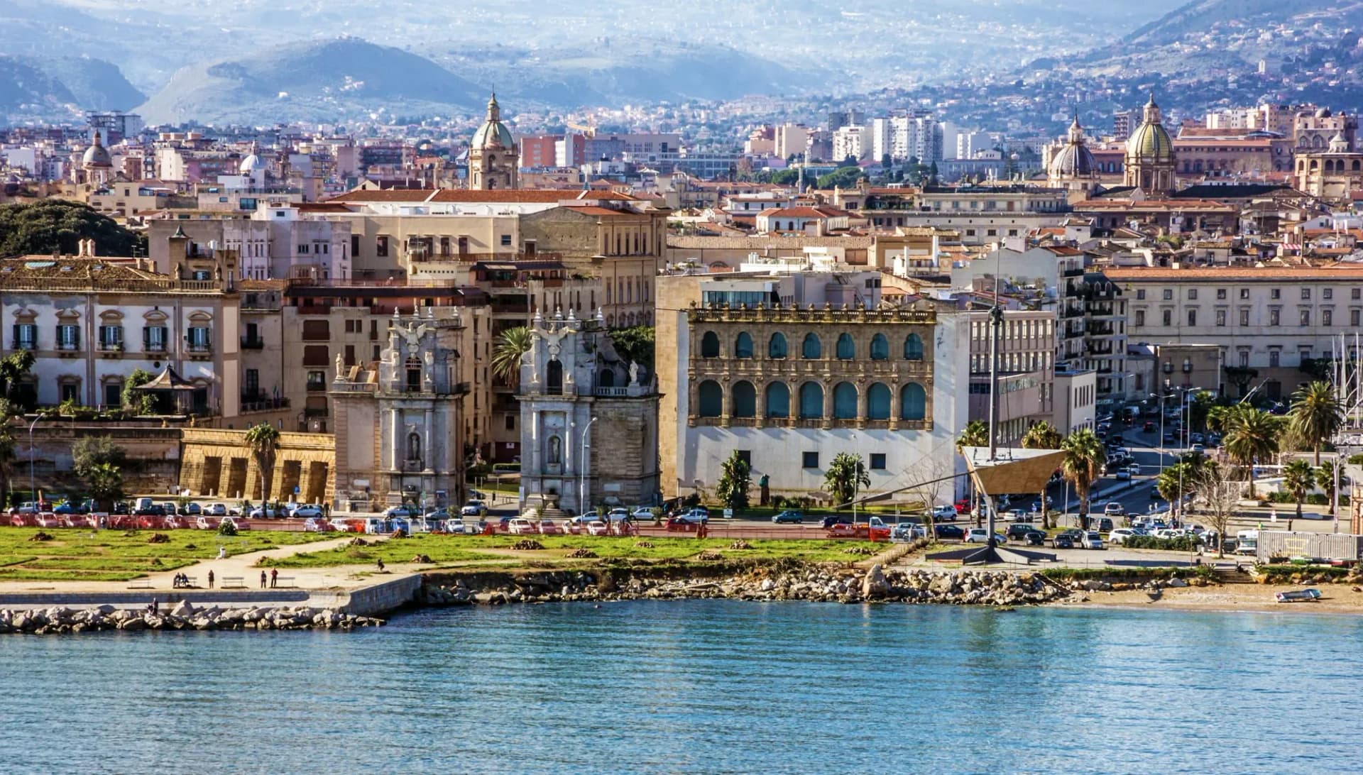 City skyline of Palermo with historic buildings, green space, and blue sea in the foreground.