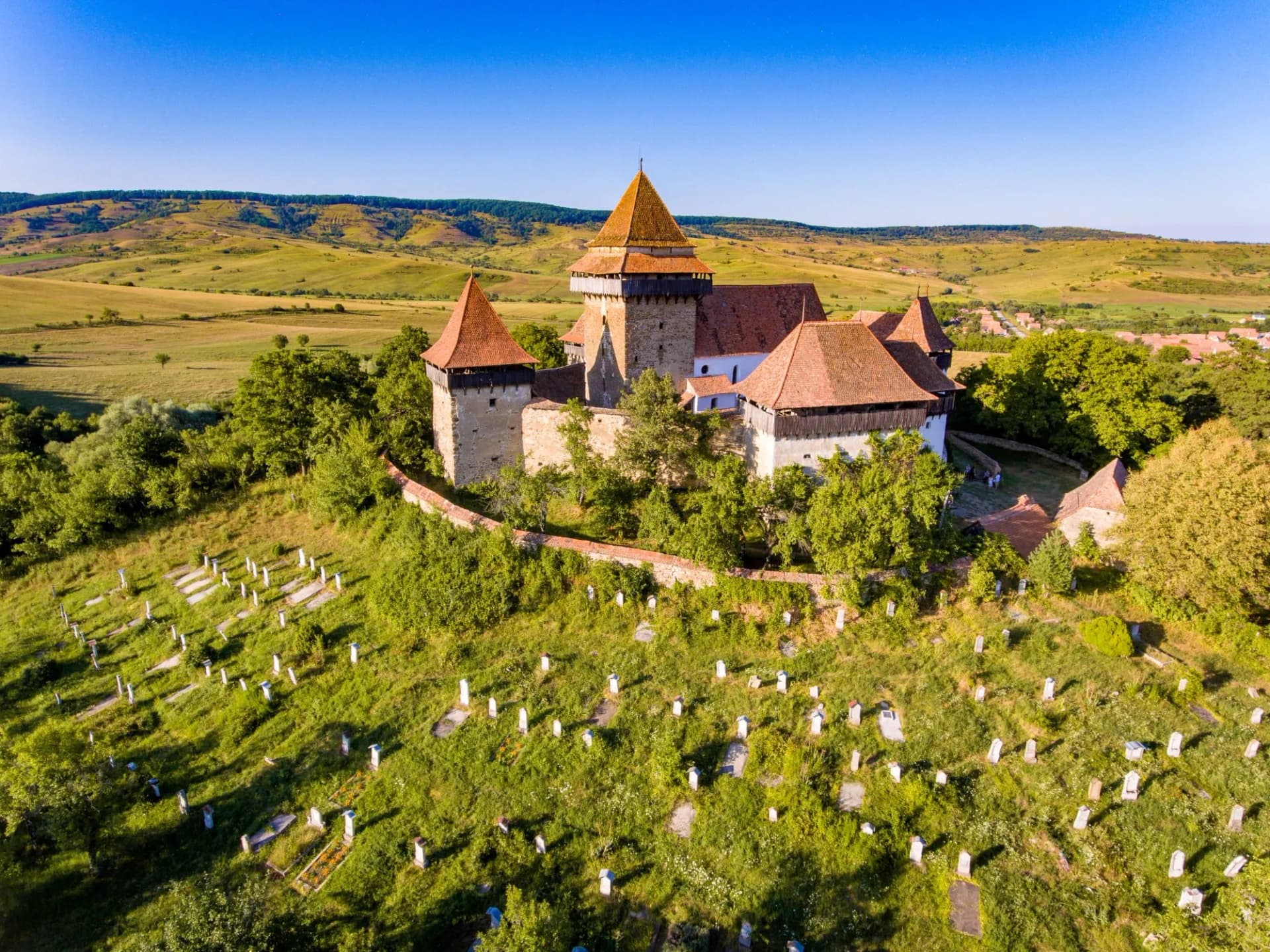 Viscri fortified church with cemetery on grassy hill, surrounded by rolling green countryside.