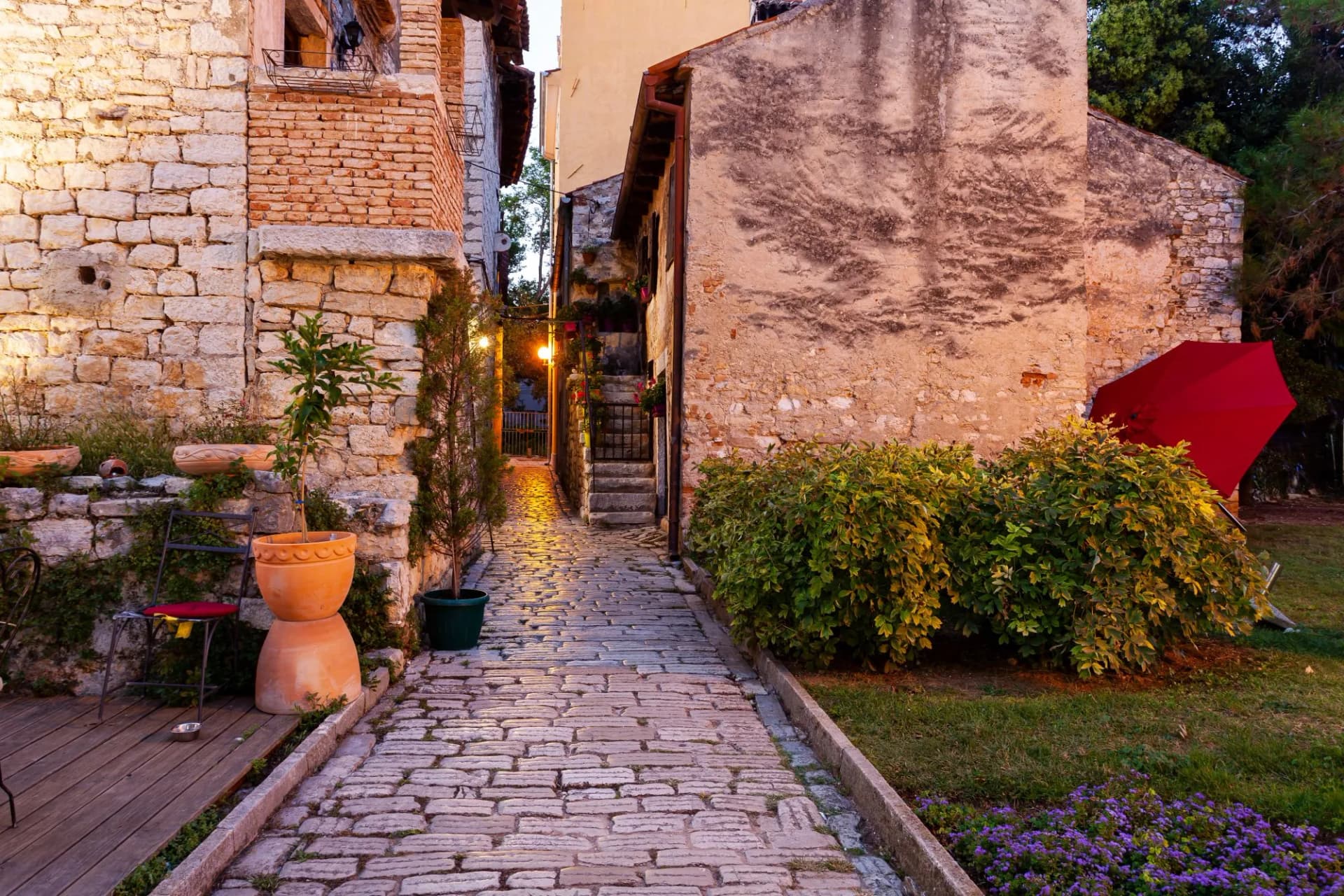 Cobblestone alleyway between stone buildings with potted plants and a red umbrella in Poreč.