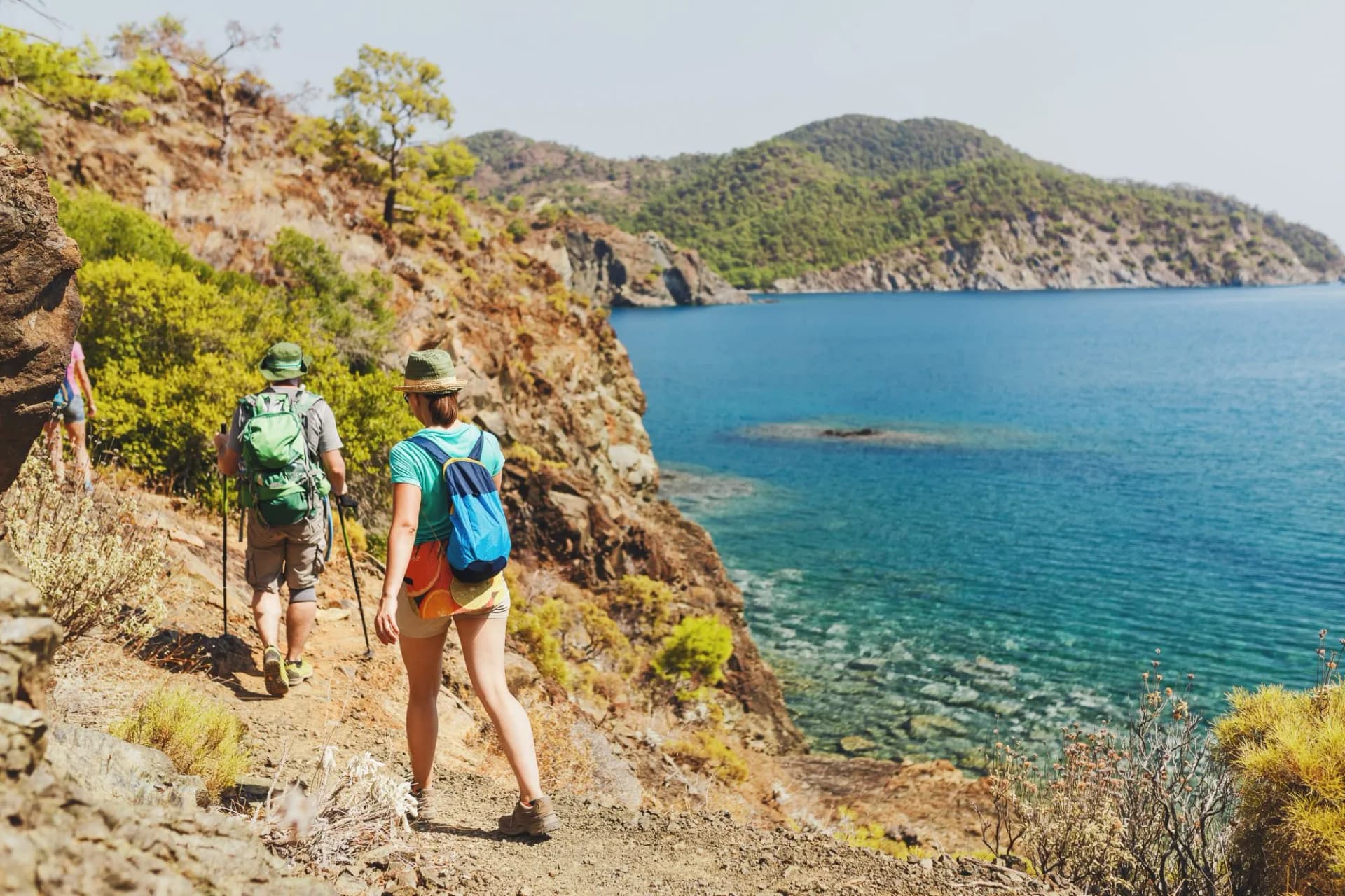 Hikers with backpacks walking on a dry coastal trail overlooking turquoise Mediterranean sea and green hills.