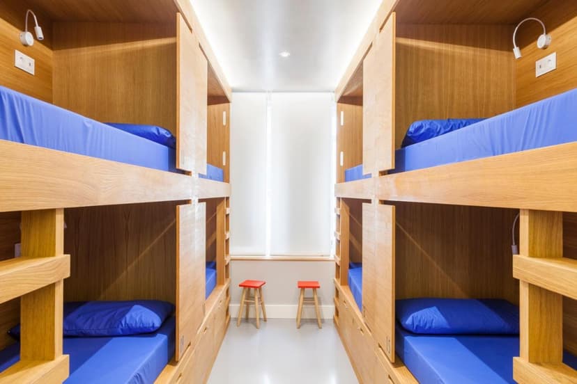 Wooden bunk beds with blue mattresses in a hostel room with a central window and two red stools.