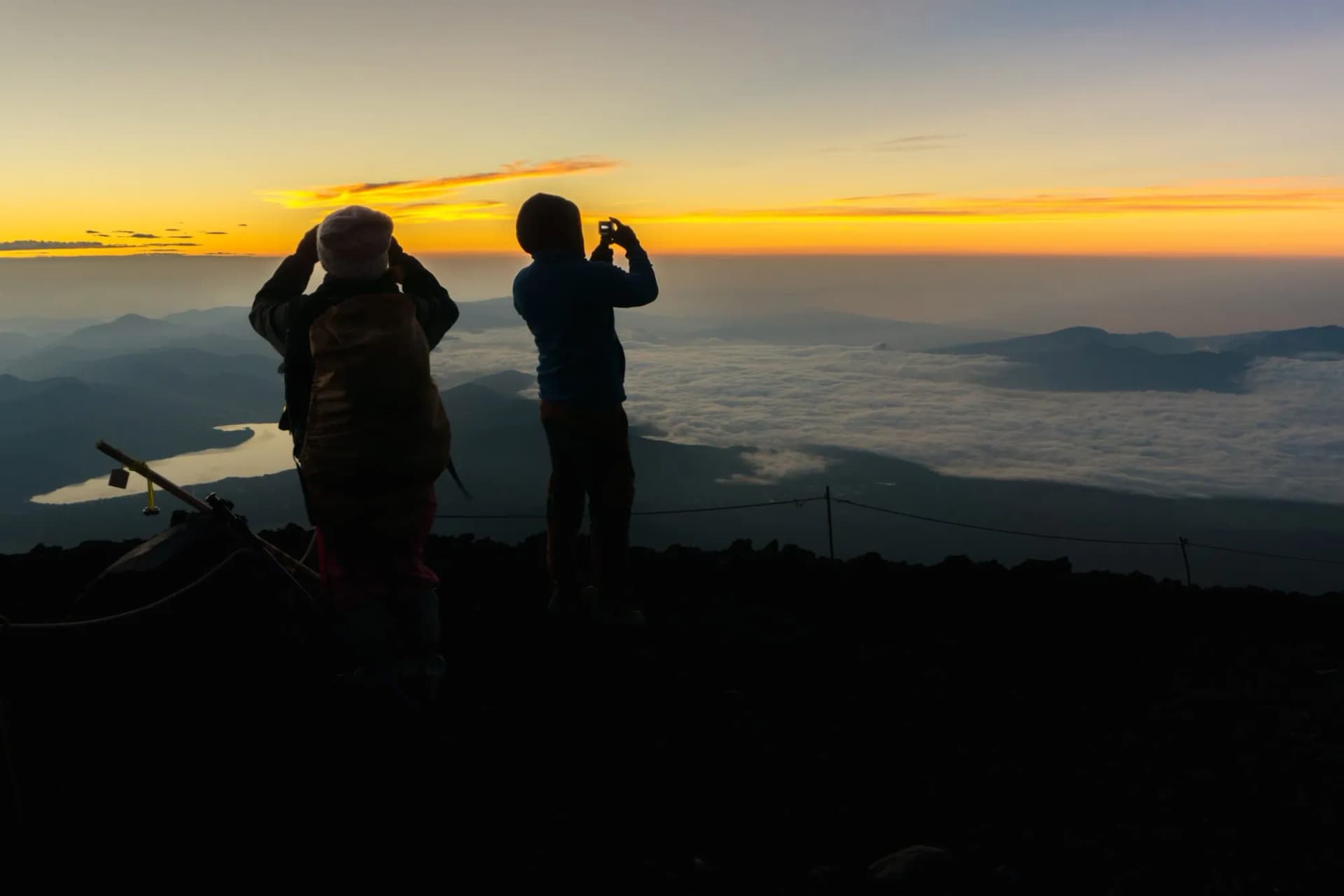 Hikers silhouetted watching sunrise above clouds with lake visible, Mt Fuji area.
