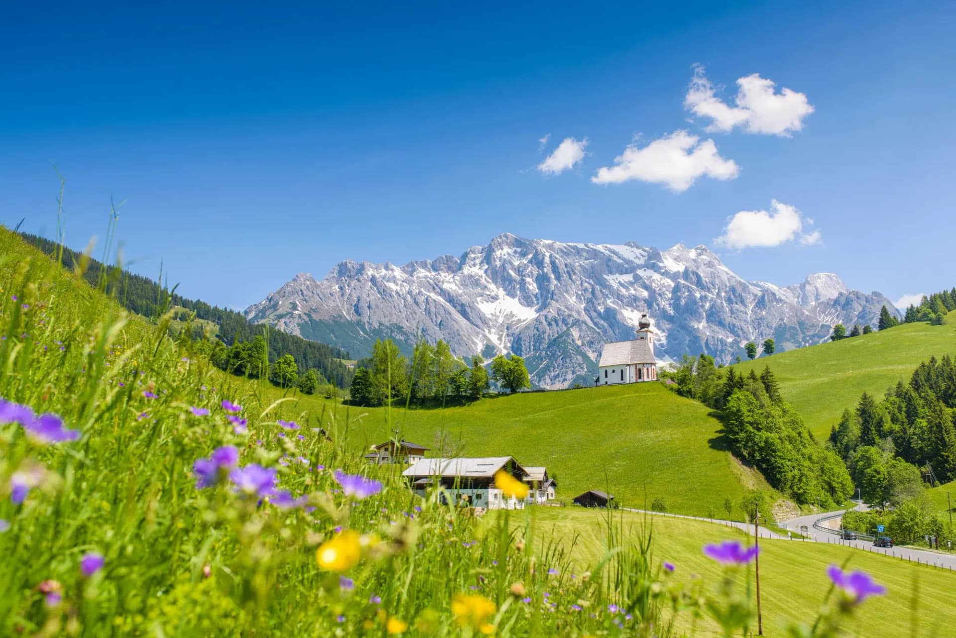 Alpine village Dienten with church, green meadows, and snow-capped mountains under blue sky.