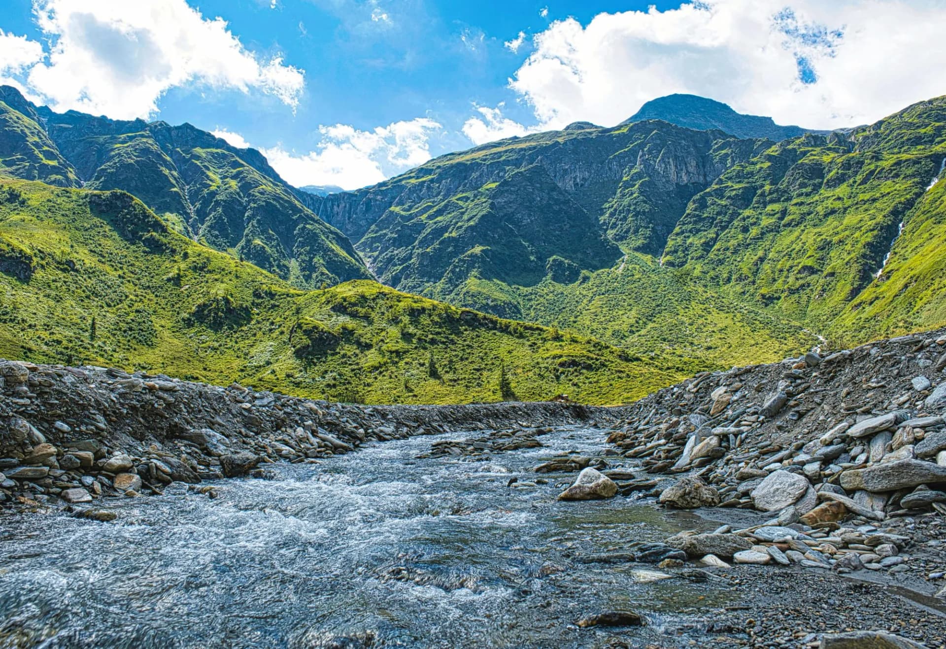 Rushing mountain stream with rocky banks flowing between steep, green slopes under a blue sky at Nassfeld-Sportgastein.