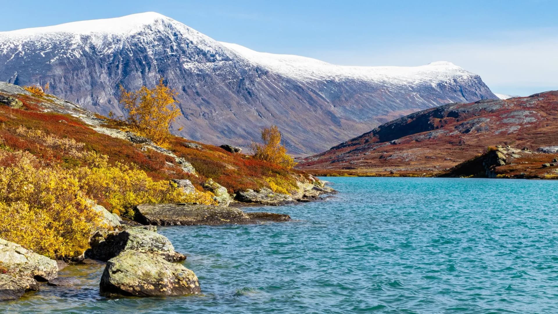 Abiskojaure Lake with turquoise water, autumn foliage, and snow-capped mountain under blue sky.