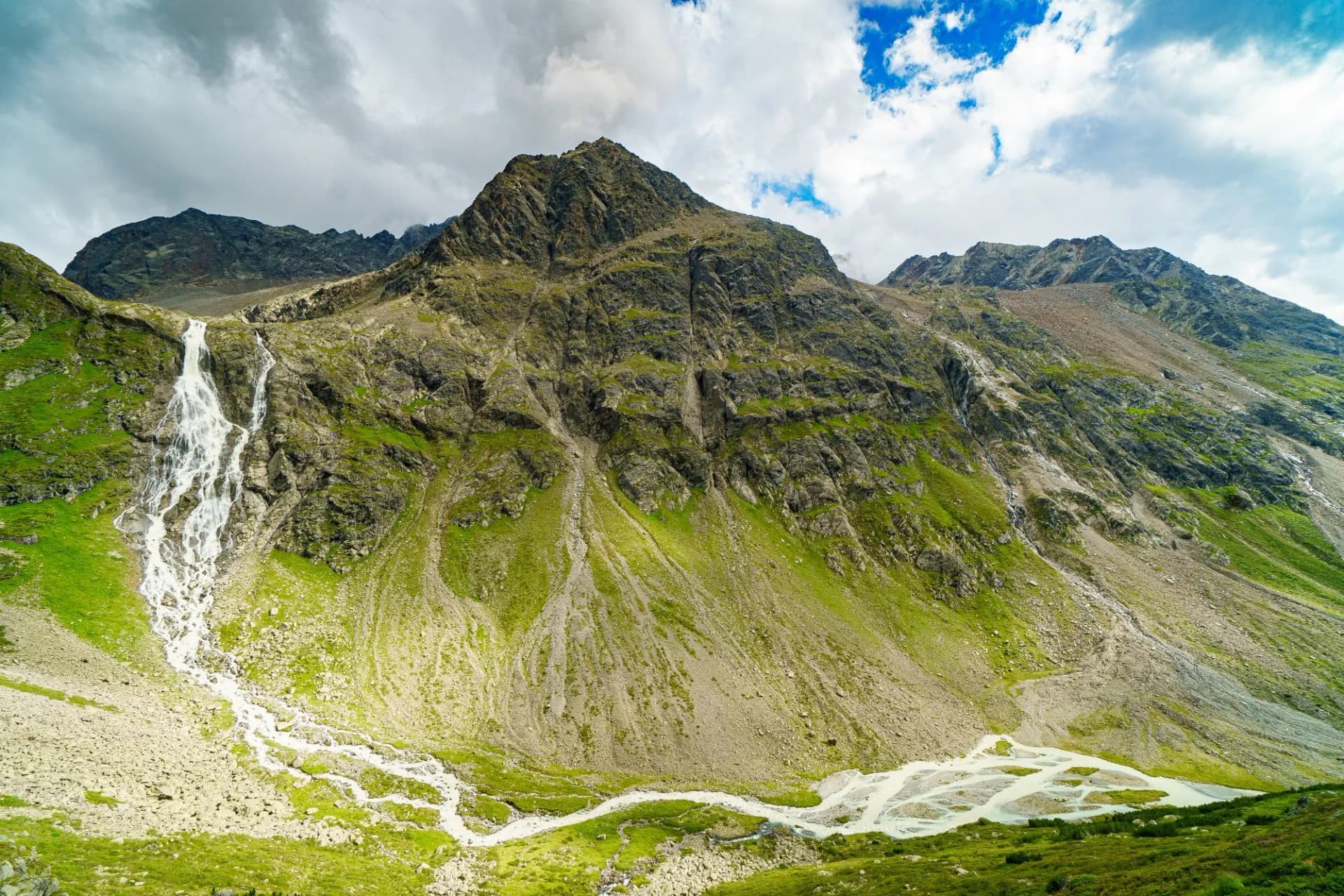 Waterfall cascading down green and rocky mountainside in Otztal under cloudy sky.
