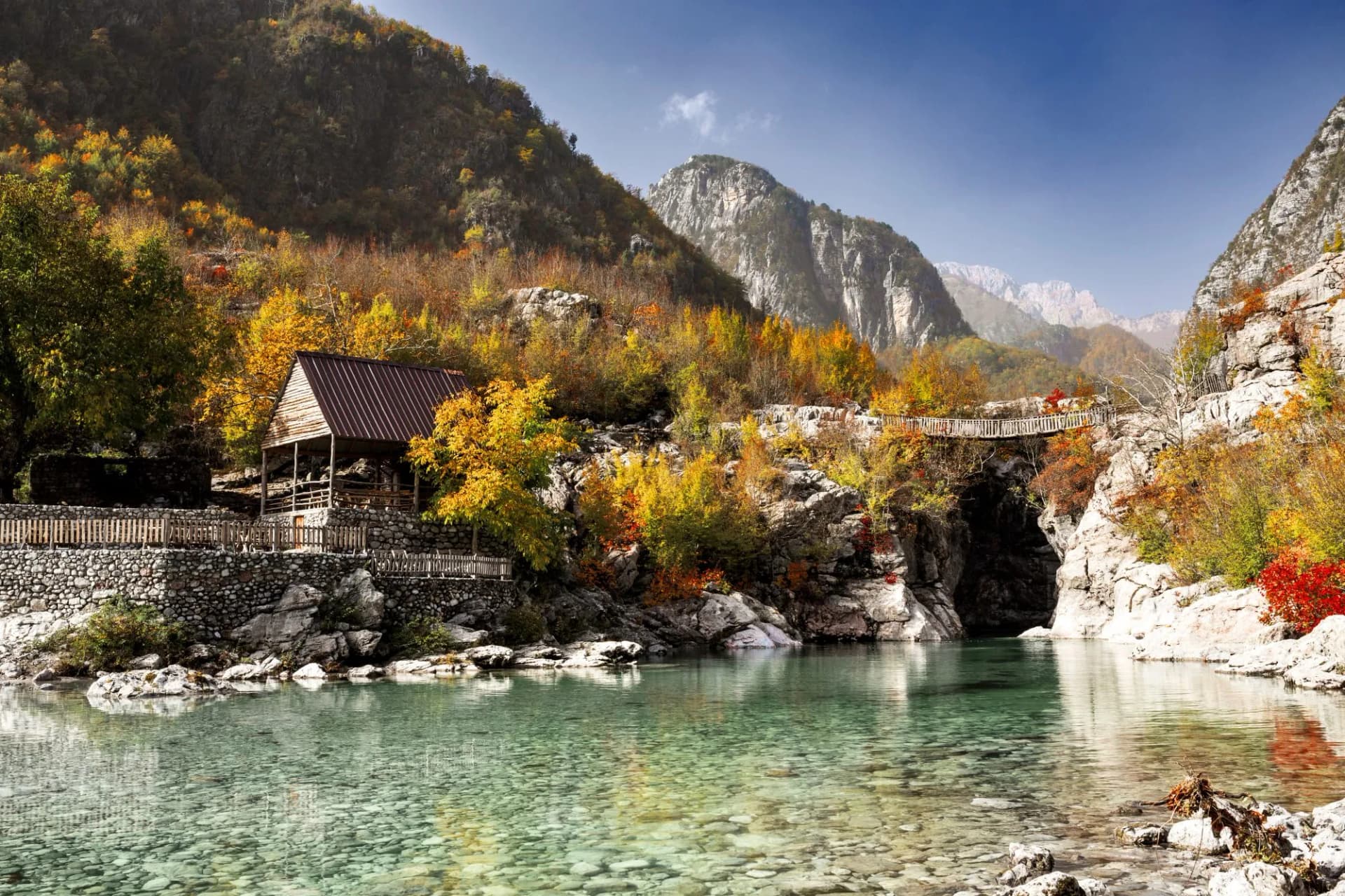 Clear river water flows past a wooden structure and footbridge surrounded by mountains with autumn foliage.