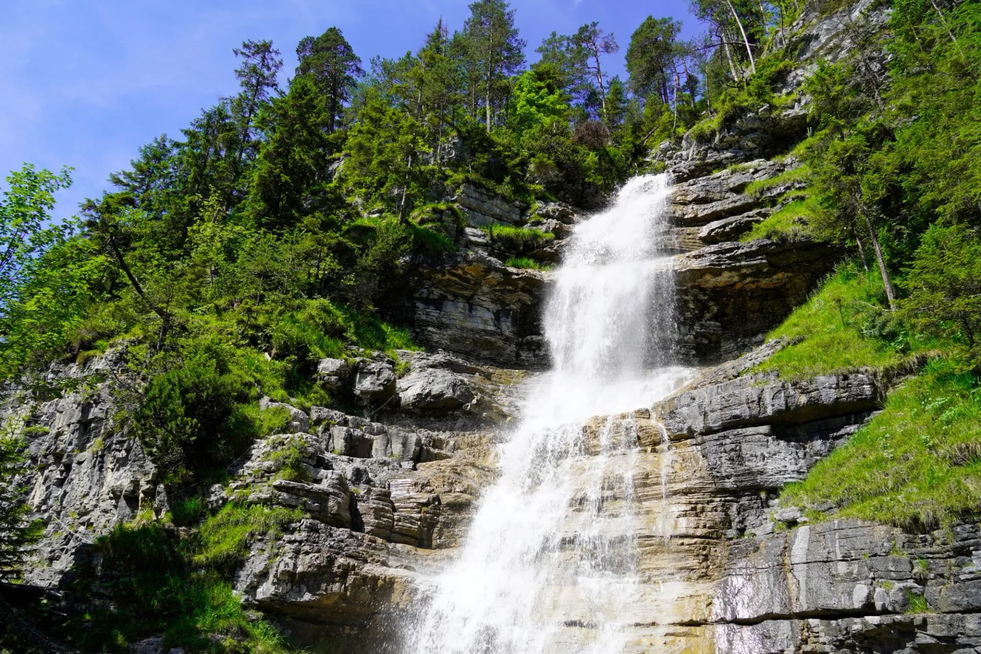 Haselgehr waterfall cascading down layered rock cliffs surrounded by green forest under a blue sky in Tyrol.