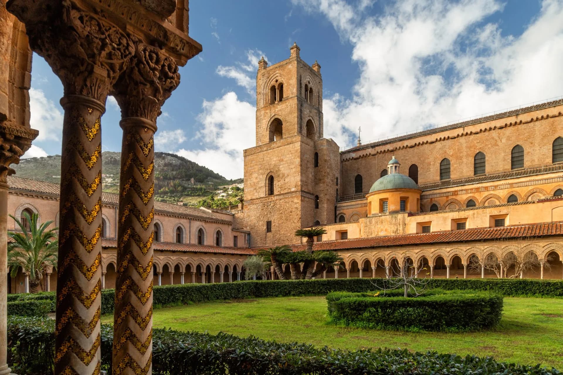 Cloister courtyard with ornate columns, bell tower, and dome of the Cathedral of Monreale.