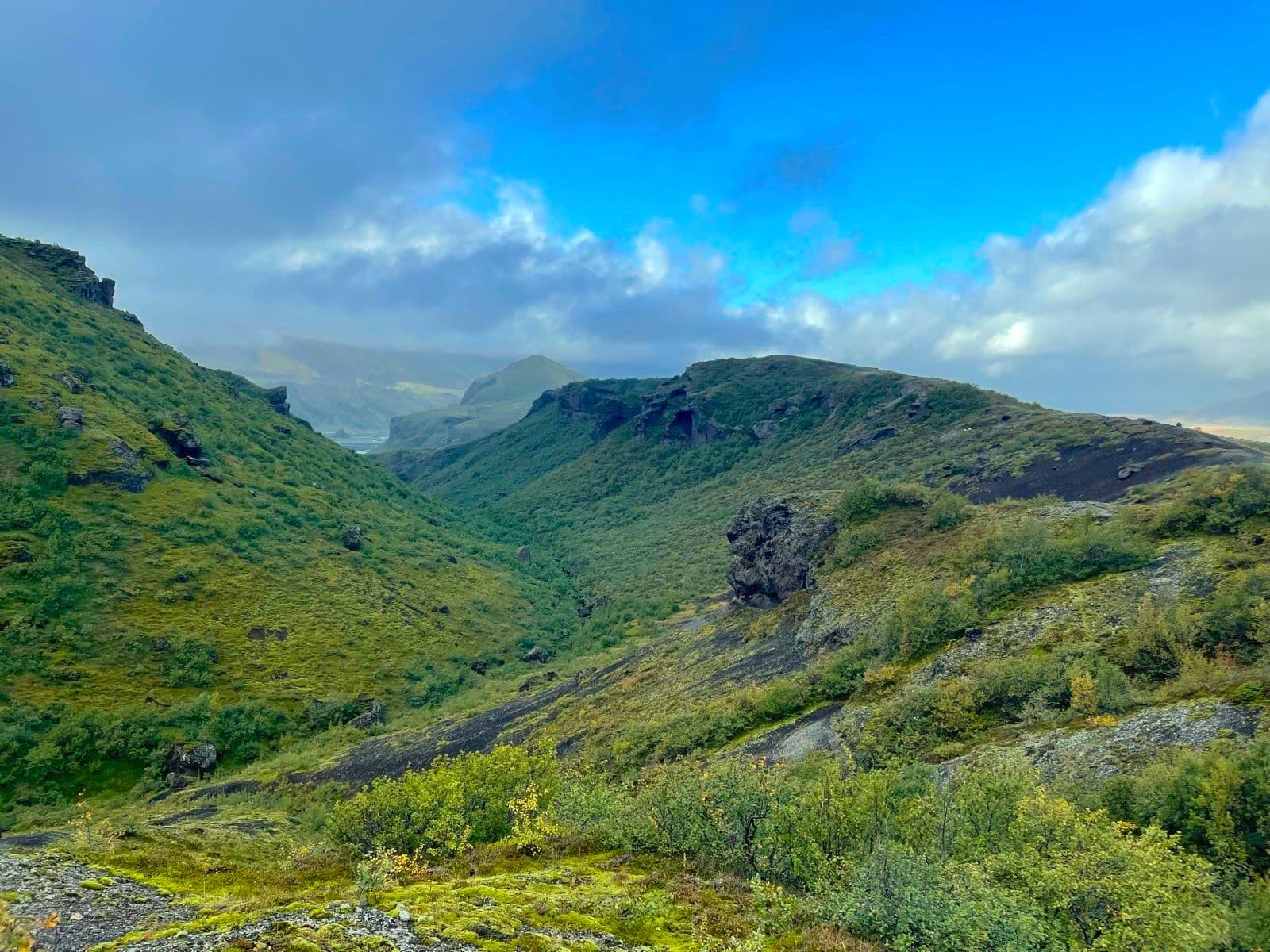 Hiking trail overlooking a lush green valley with steep moss-covered hills under a blue sky.