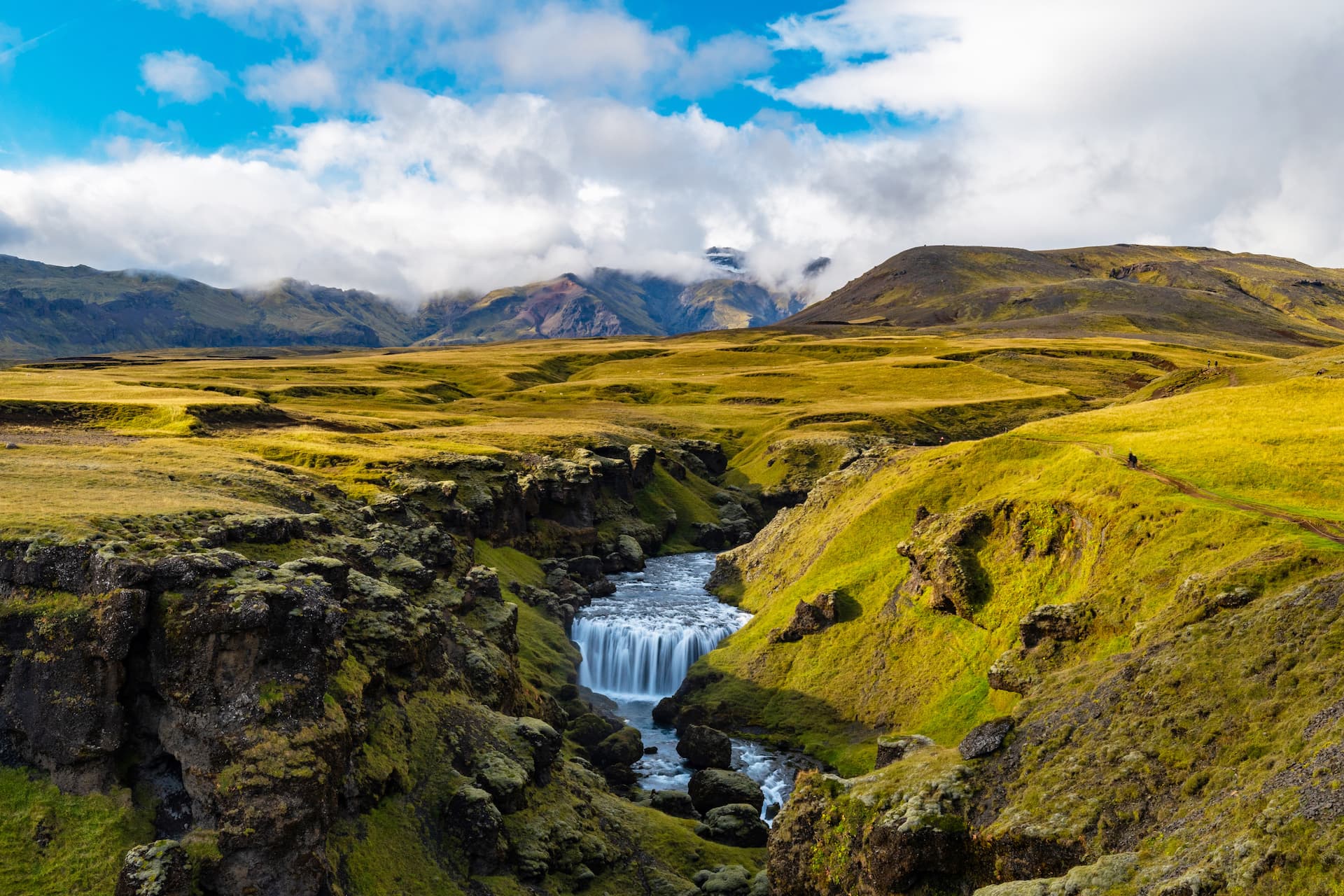Small waterfall in a canyon with bright green mossy banks and distant mountains under a cloudy blue sky.