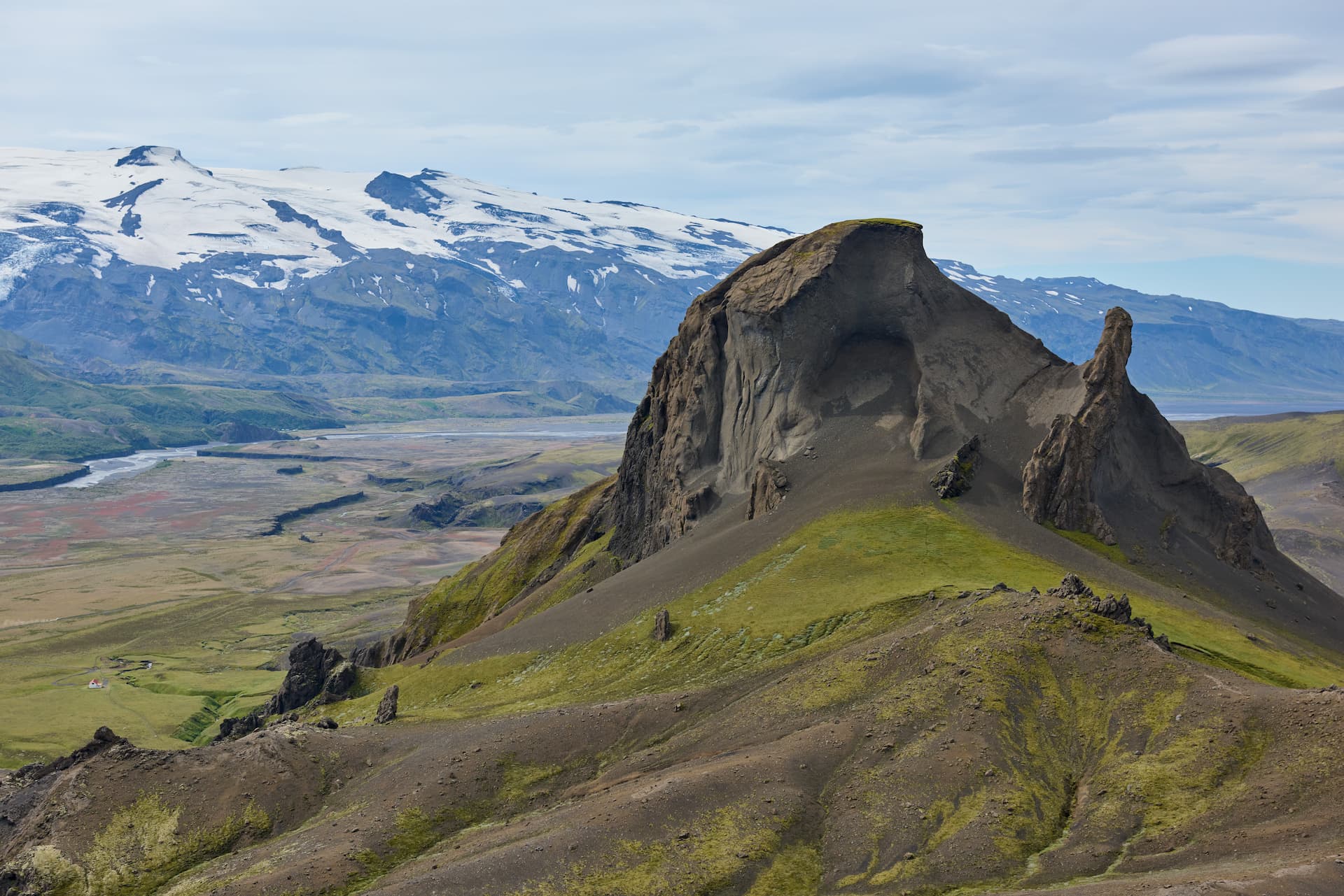 Jagged volcanic peak with green slopes overlooking a glacial river valley and snow-capped mountains.
