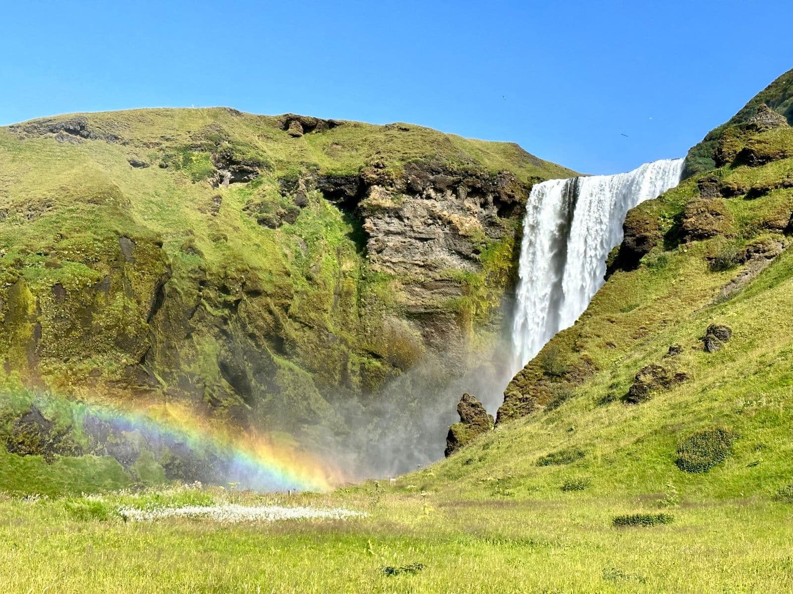 Waterfall at Fimmvörðuháls near Skógar with a rainbow over green hills under a clear blue sky.