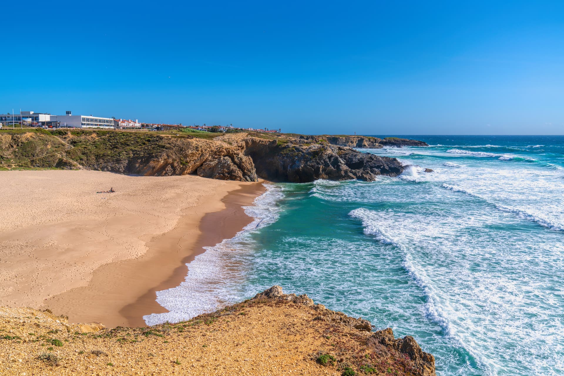 Sandy Praia Grande beach with cliffs and turquoise waves under a clear blue sky in Porto Covo.