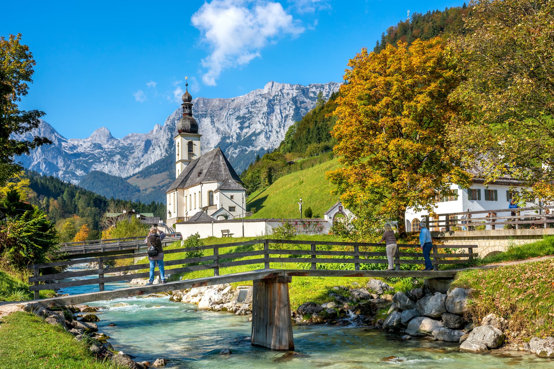 Church near Berchtesgaden with autumn trees and hikers crossing a wooden bridge over a stream.