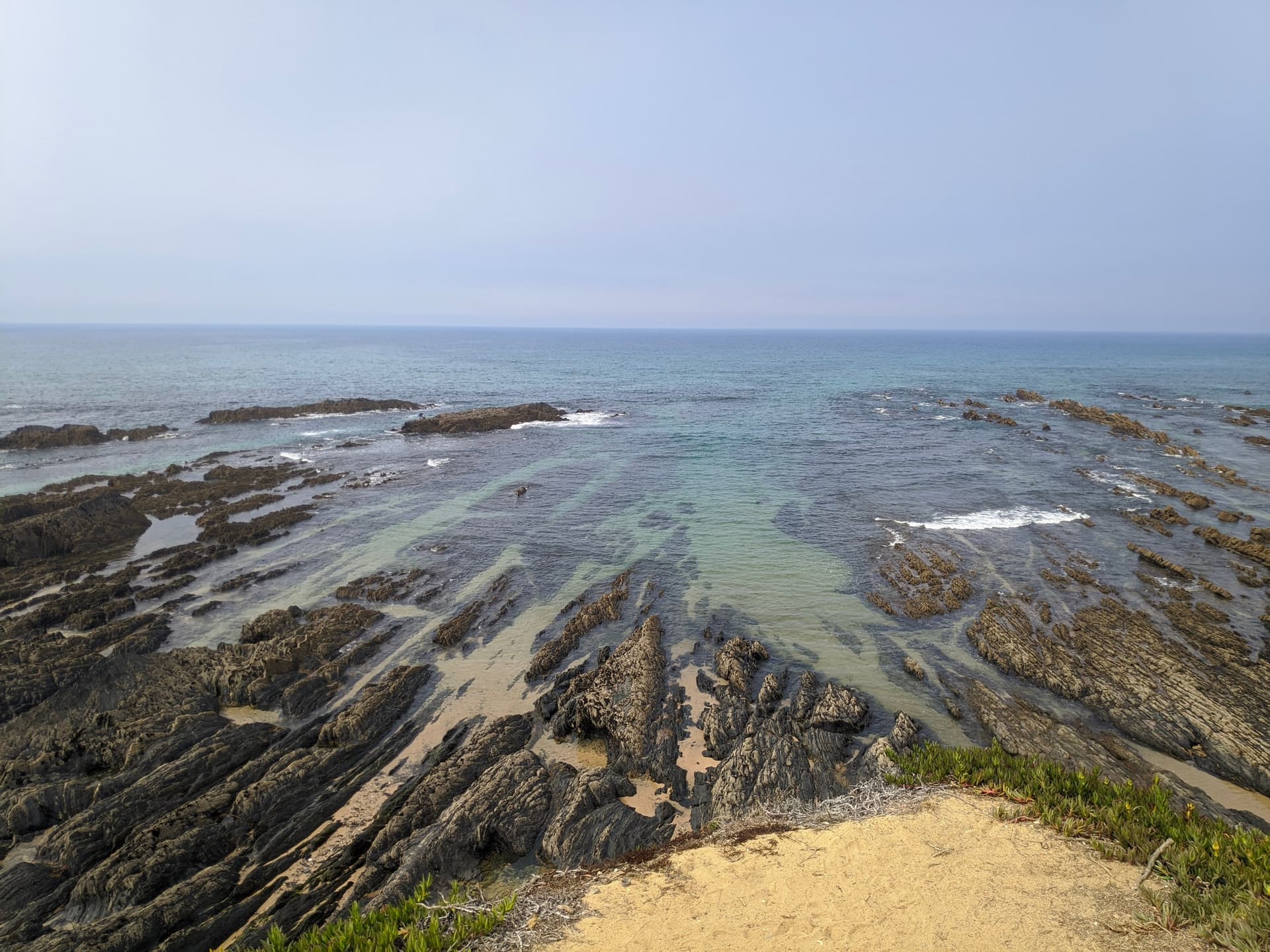 Exposed stratified rocks at low tide near Almograve beach with clear turquoise ocean water.