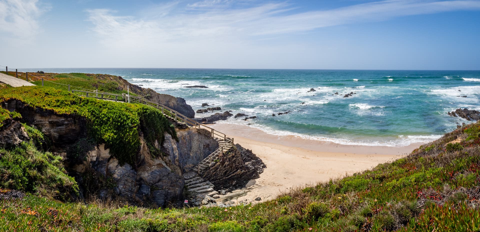 Wooden steps down a vegetated cliff to a sandy beach and turquoise ocean at Almograve.