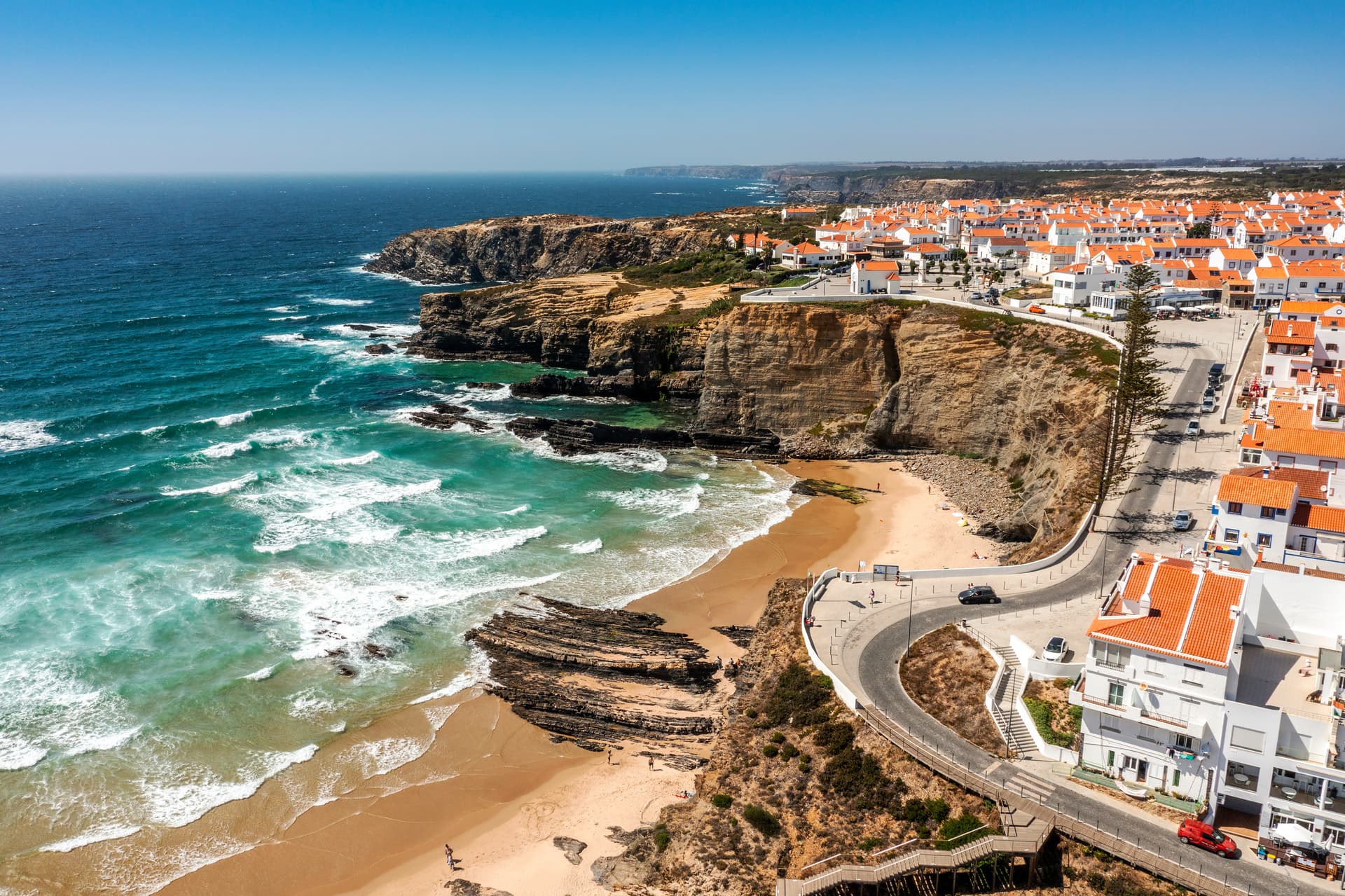 Aerial view of Zambujeira do Mar coastal town with white buildings and orange roofs above a sandy beach.