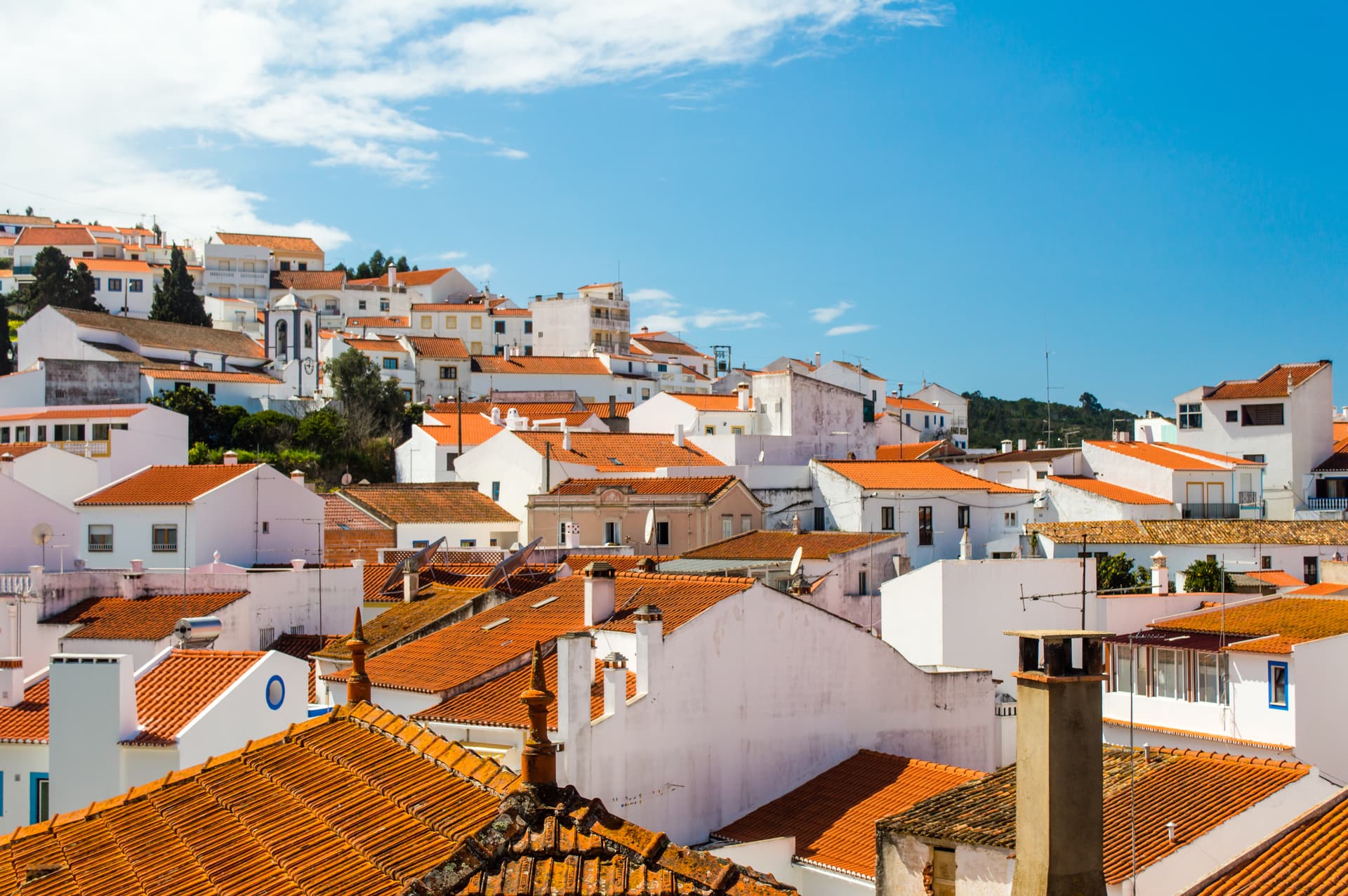 White buildings with orange terracotta roofs clustered on a hillside under a bright blue sky in Odeceixe, Portugal.