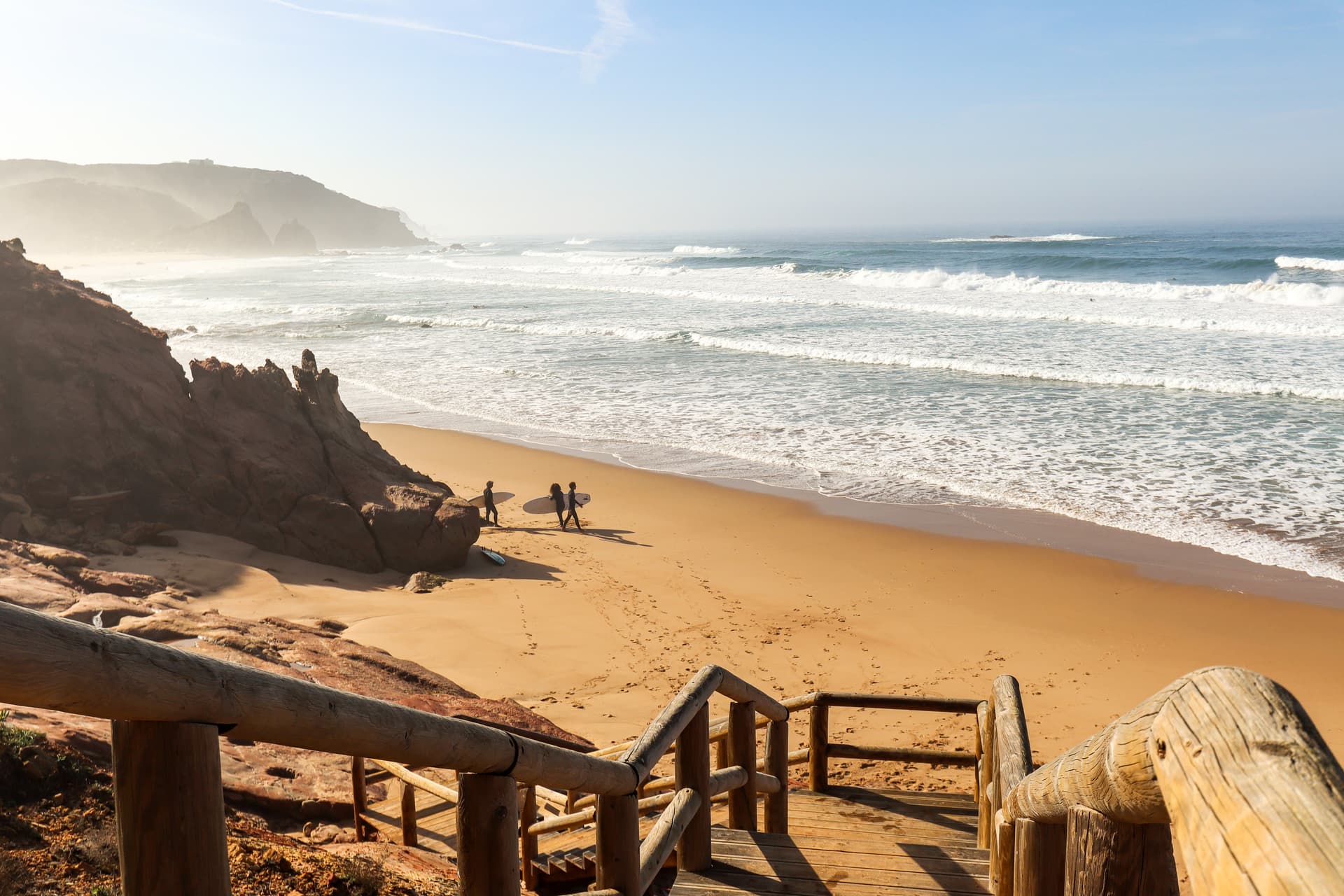 Surfers walking on Arrifana Beach with wooden stairs overlooking the Atlantic waves in Portugal.