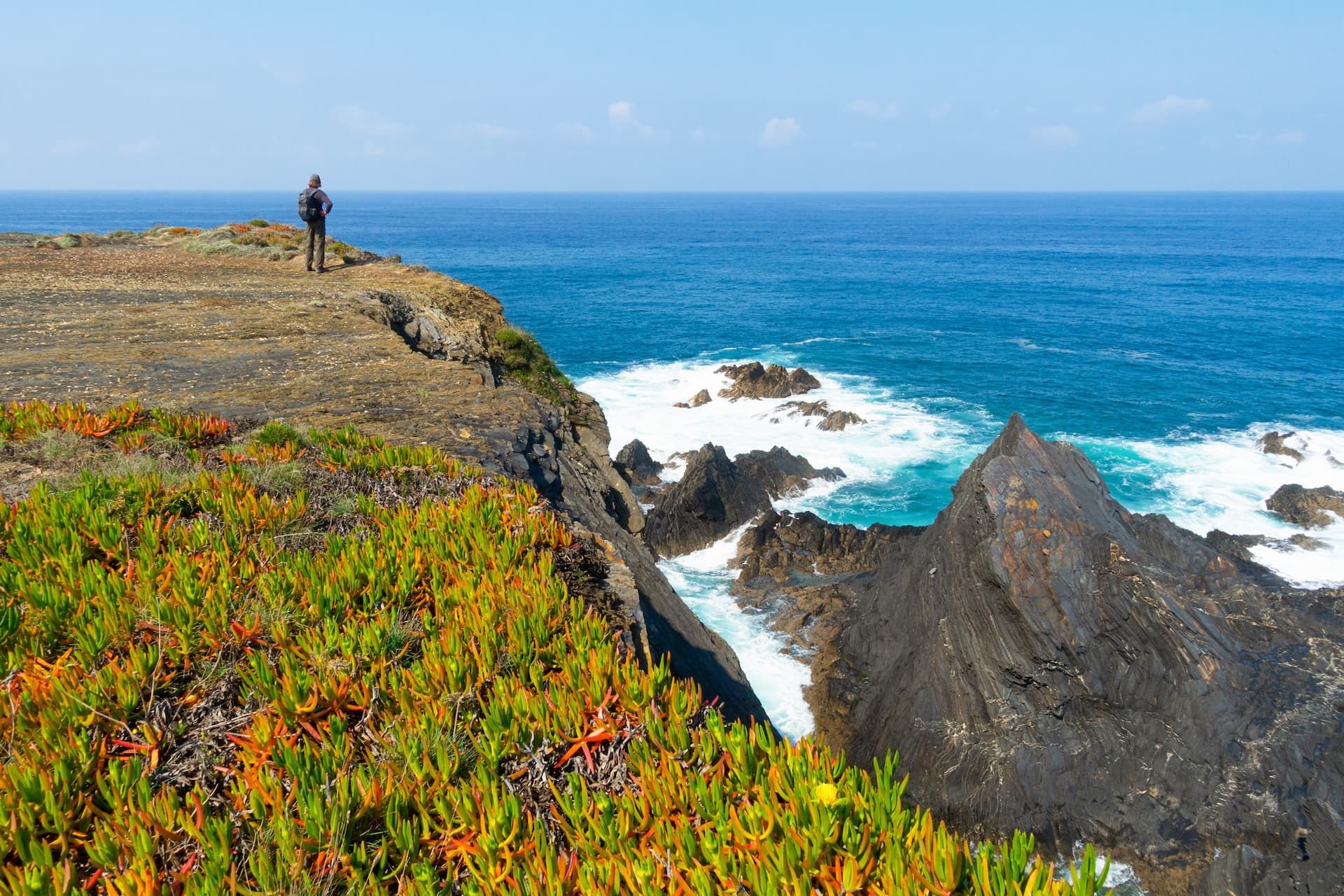 Hiker overlooking rugged coastline, sea cliffs, and turquoise water near Almograve.