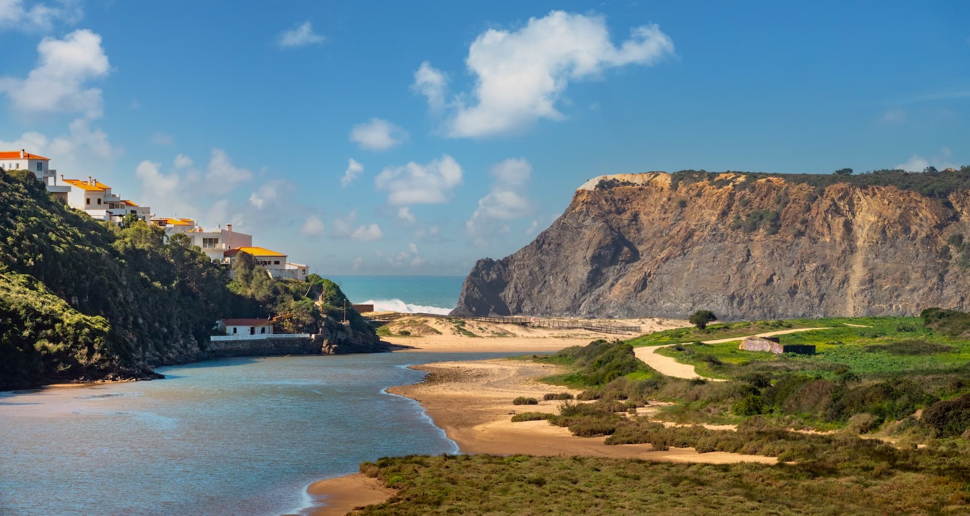 River estuary meeting the ocean at Praia de Odeceixe on the Rota Vicentina, with cliffs and white houses.