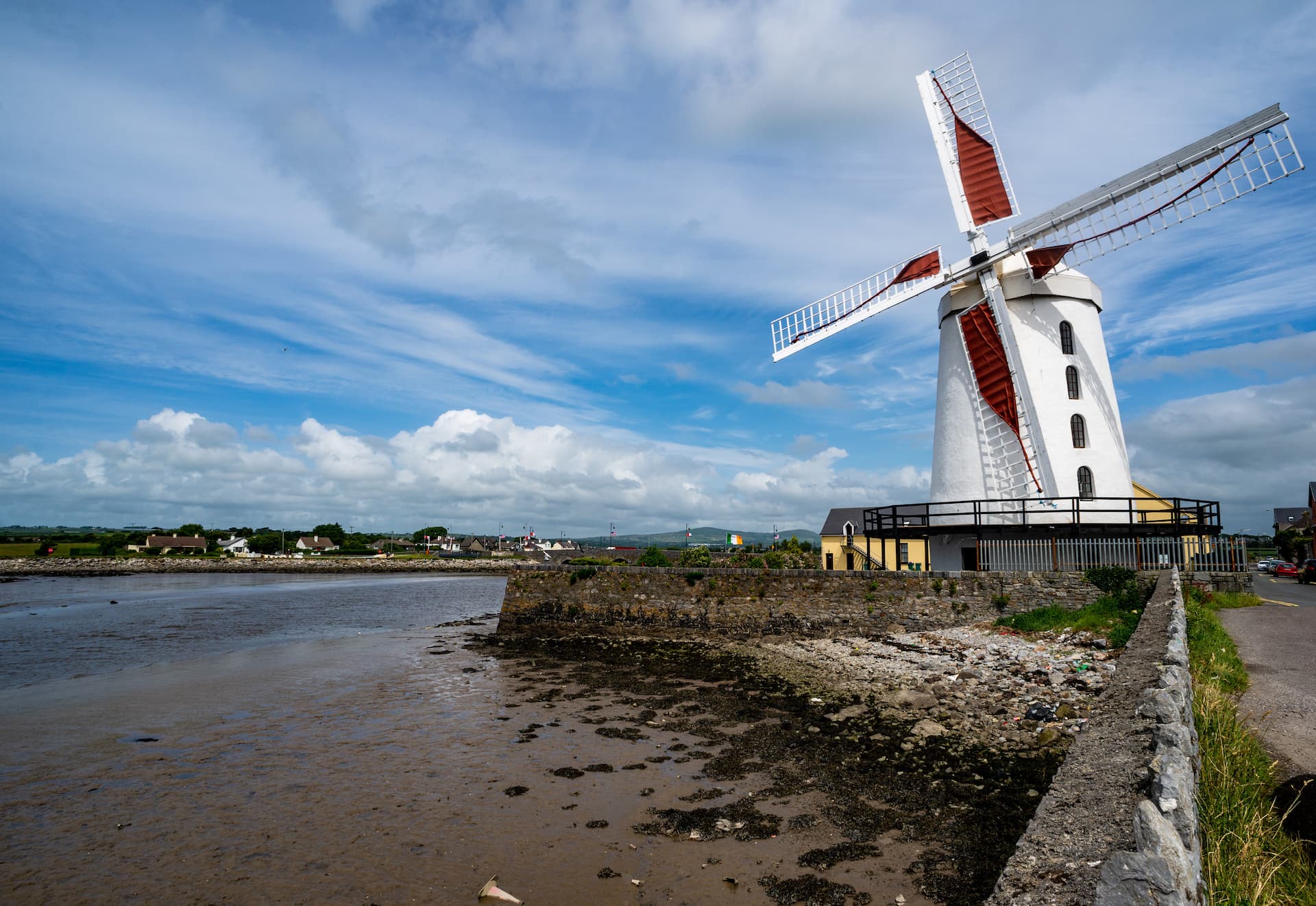 White Tralee windmill with red sails beside muddy tidal flats under a cloudy blue sky.