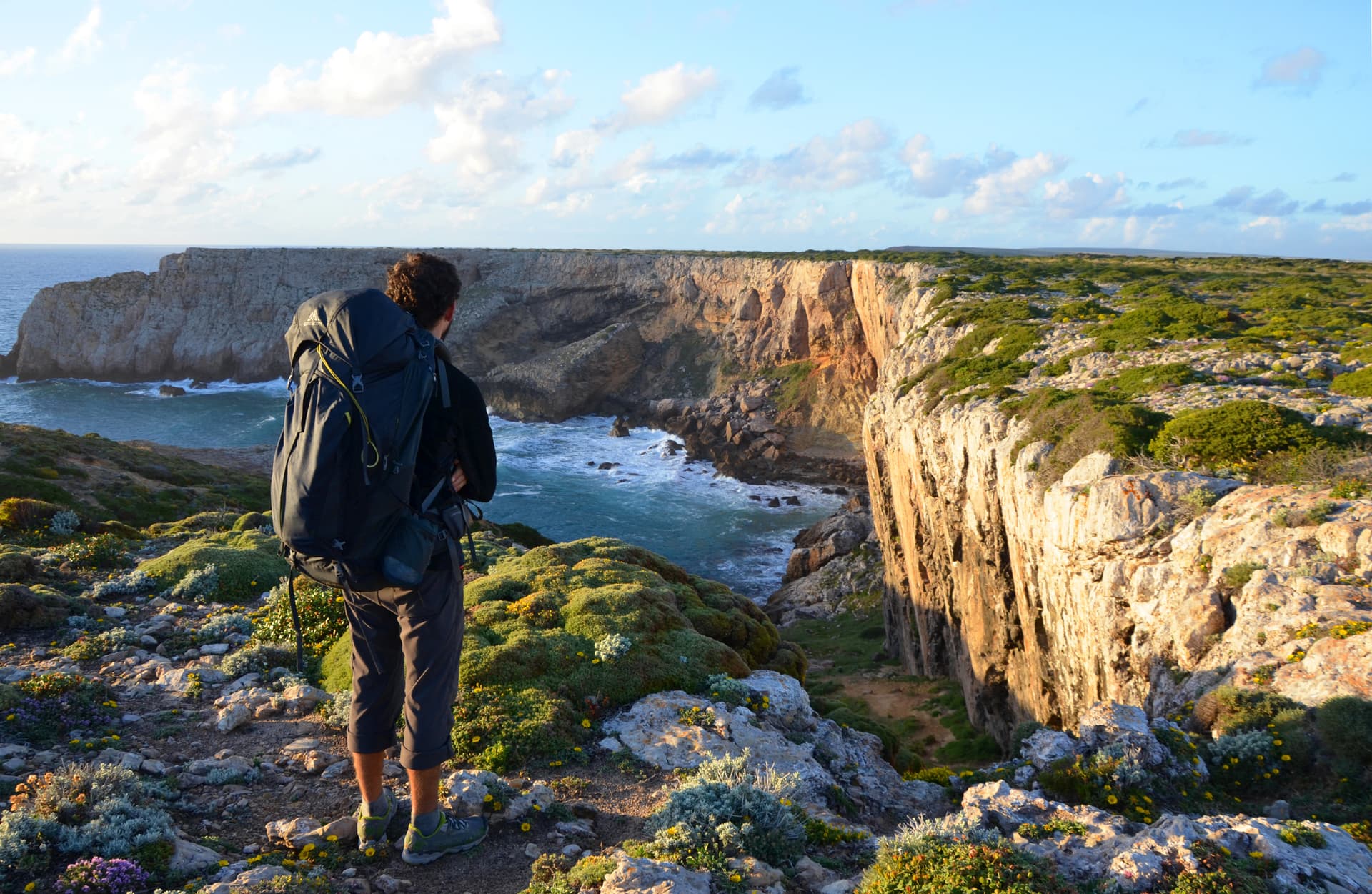 Hiker with backpack overlooking rugged cliffs and churning sea water on sunny day