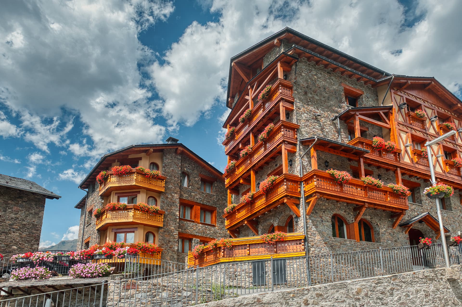 Stone and wood alpine buildings with balconies overflowing with red and pink flowers under a cloudy blue sky.