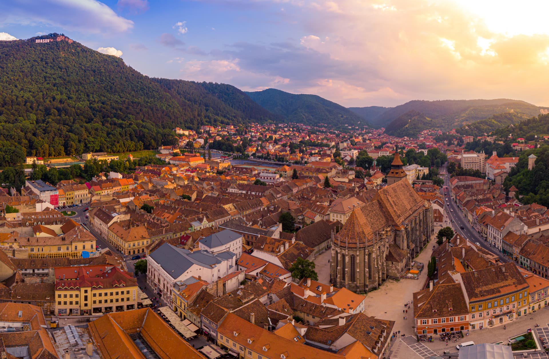 Aerial view of Brasov city rooftops, Black Church, and wooded mountains at sunset.