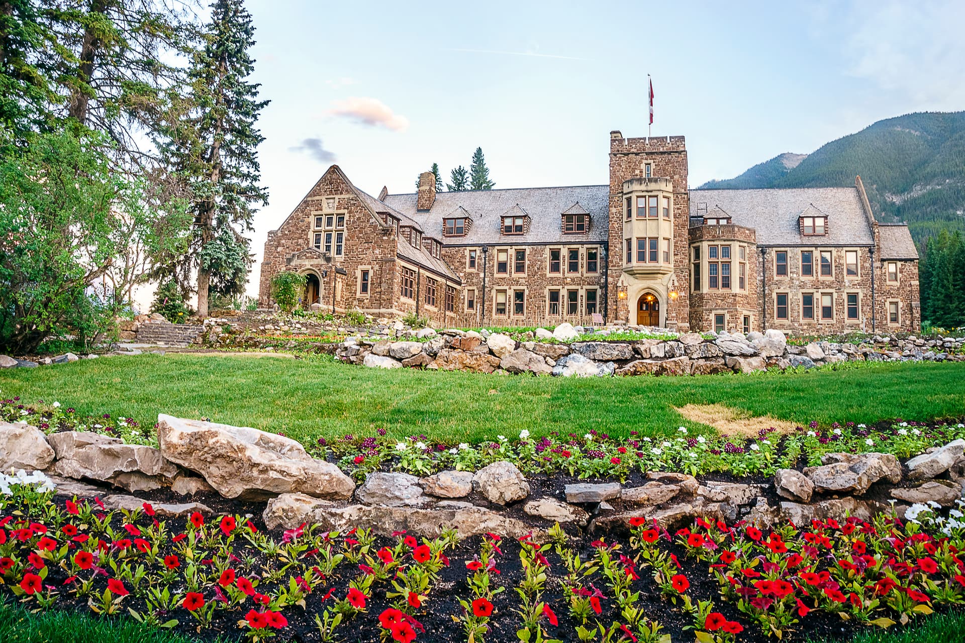 Stone castle-like building with Canadian flag, green lawn, and red flowers, set against mountains.